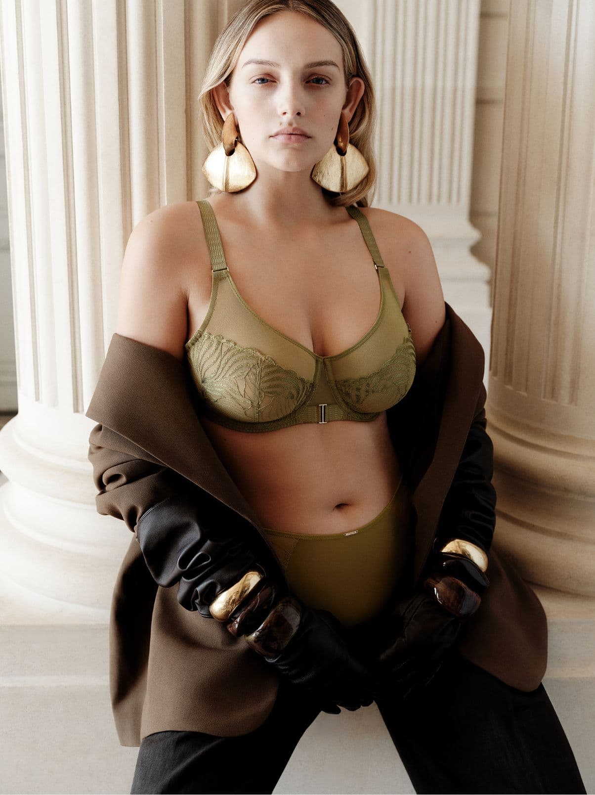 Woman in olive lingerie and oversized earrings poses confidently, wearing a brown coat and black gloves, standing by large columns.