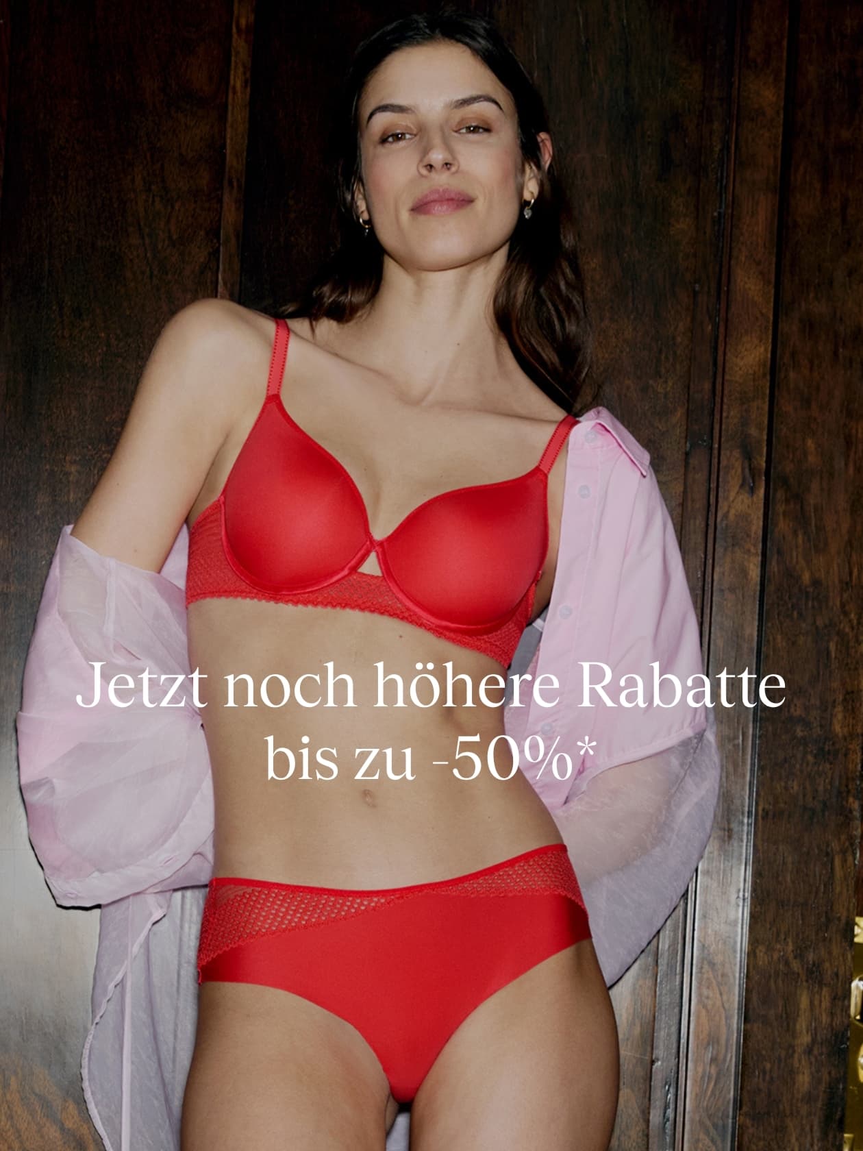 A person in red lingerie with a pink shirt draped over their arms, standing before a wooden background. German text offers up to 50% discounts.