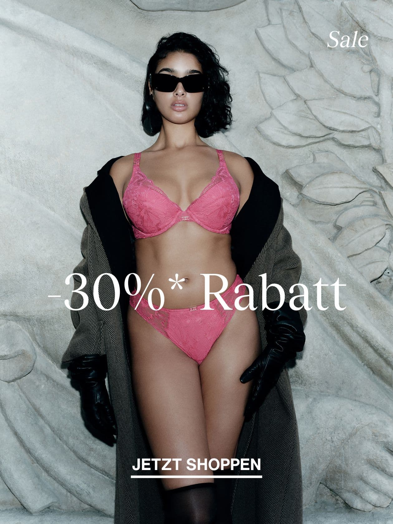 Model in pink lace bra and panties, oversized coat and black gloves, wearing sunglasses, posing against carved stone backdrop with sale text.