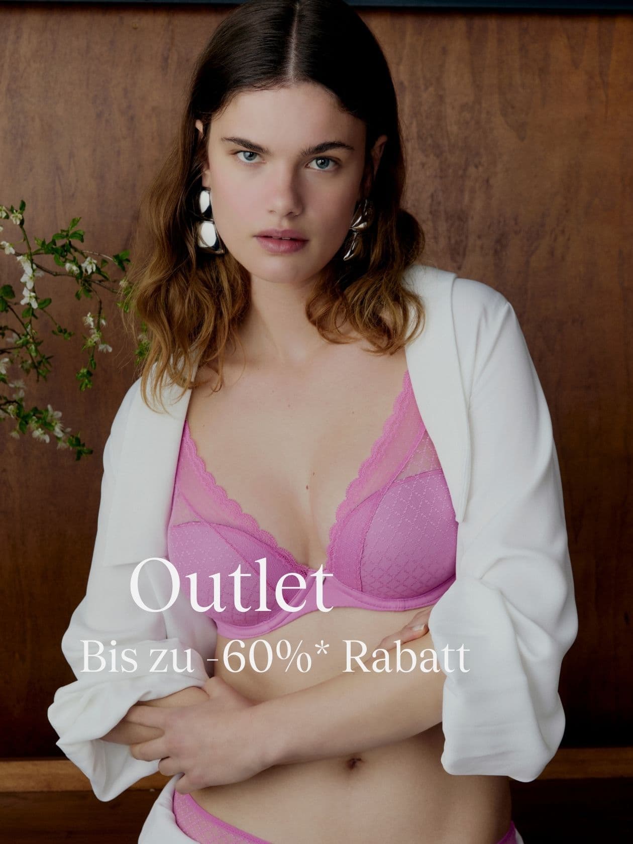 Woman in a pink lingerie set and white robe, standing in front of a wooden wall. Text on image: "Outlet Bis zu -60%* Rabatt."