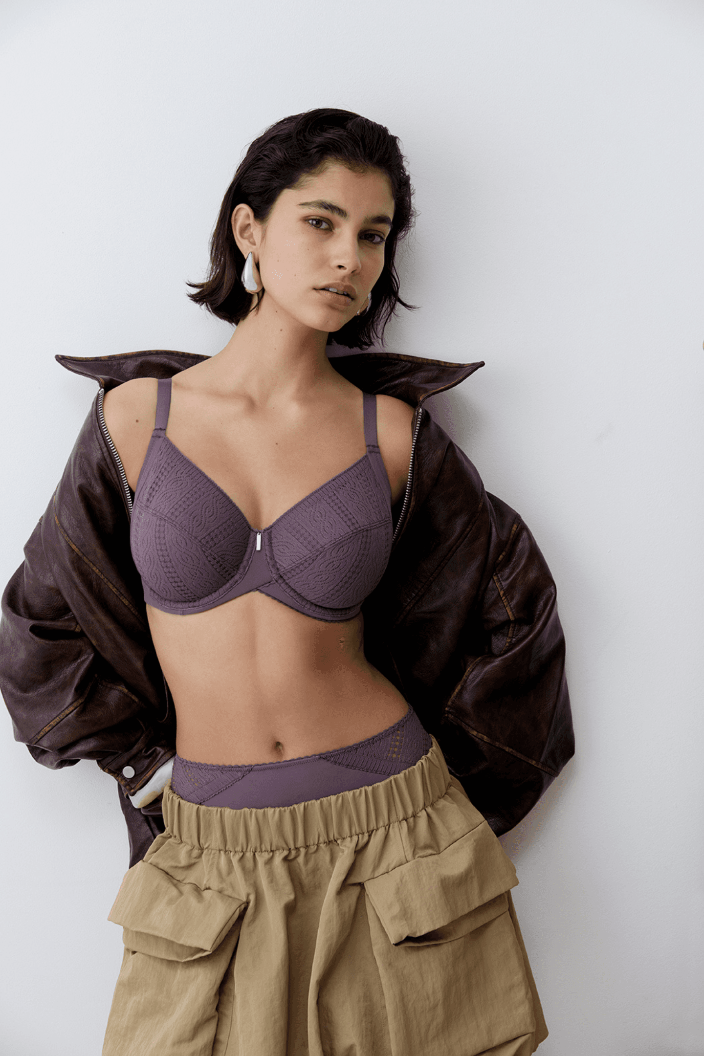 A person posing in a purple lace bra and beige skirt, with a dark leather jacket draped over their shoulders.