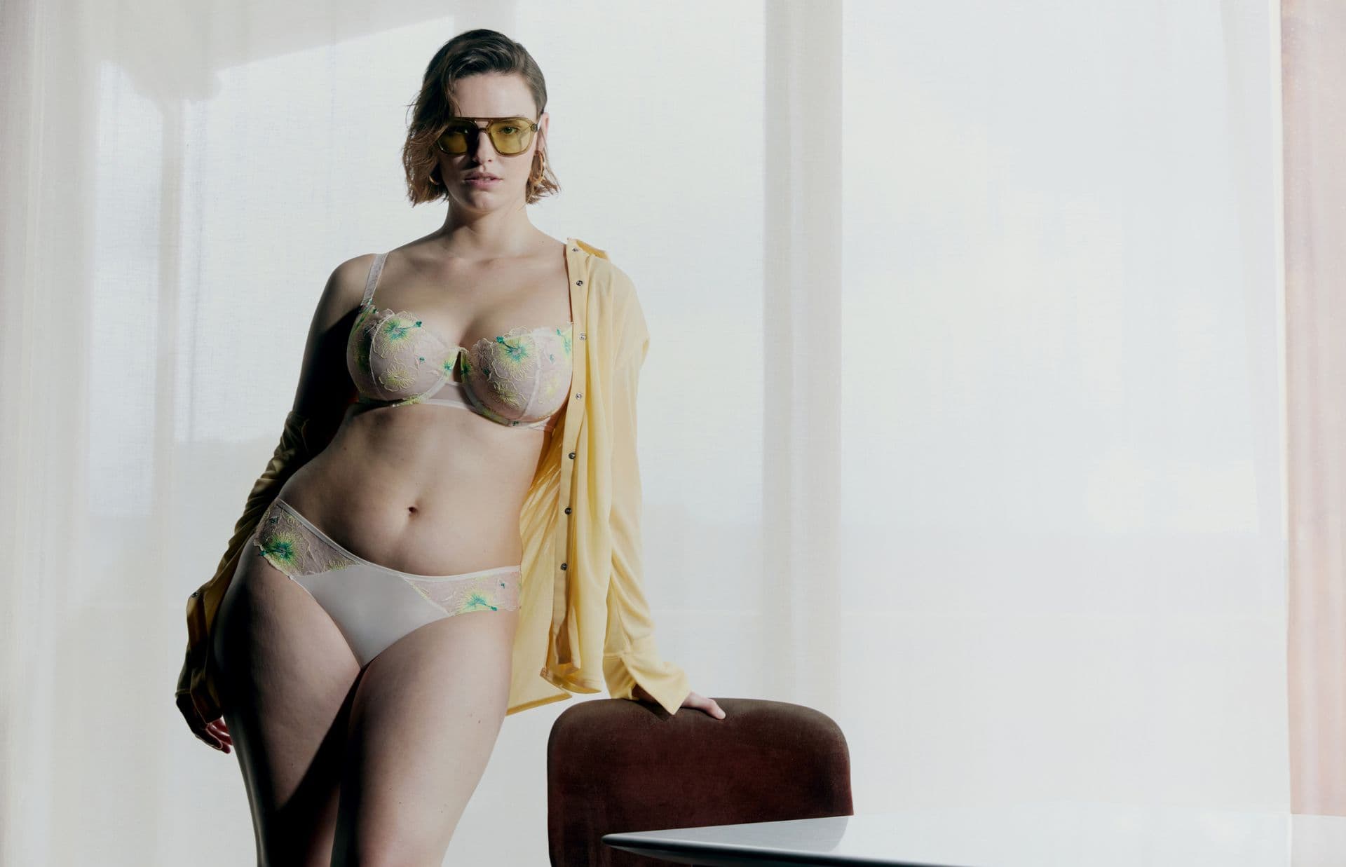 Person wearing stylish lingerie and a loose yellow shirt, standing by a chair, with sunglasses, in softly lit room.