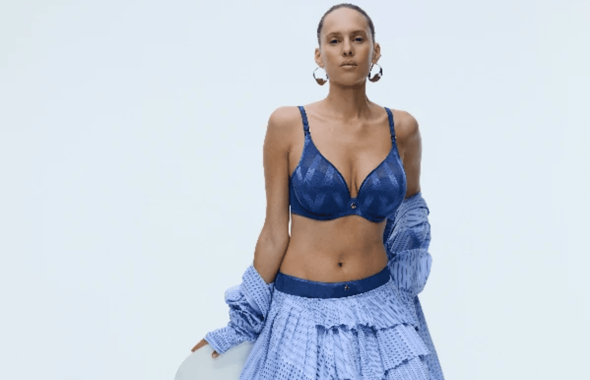 Woman wearing a blue patterned bra and skirt, with a matching open shirt, and hoop earrings, standing against a plain light background.