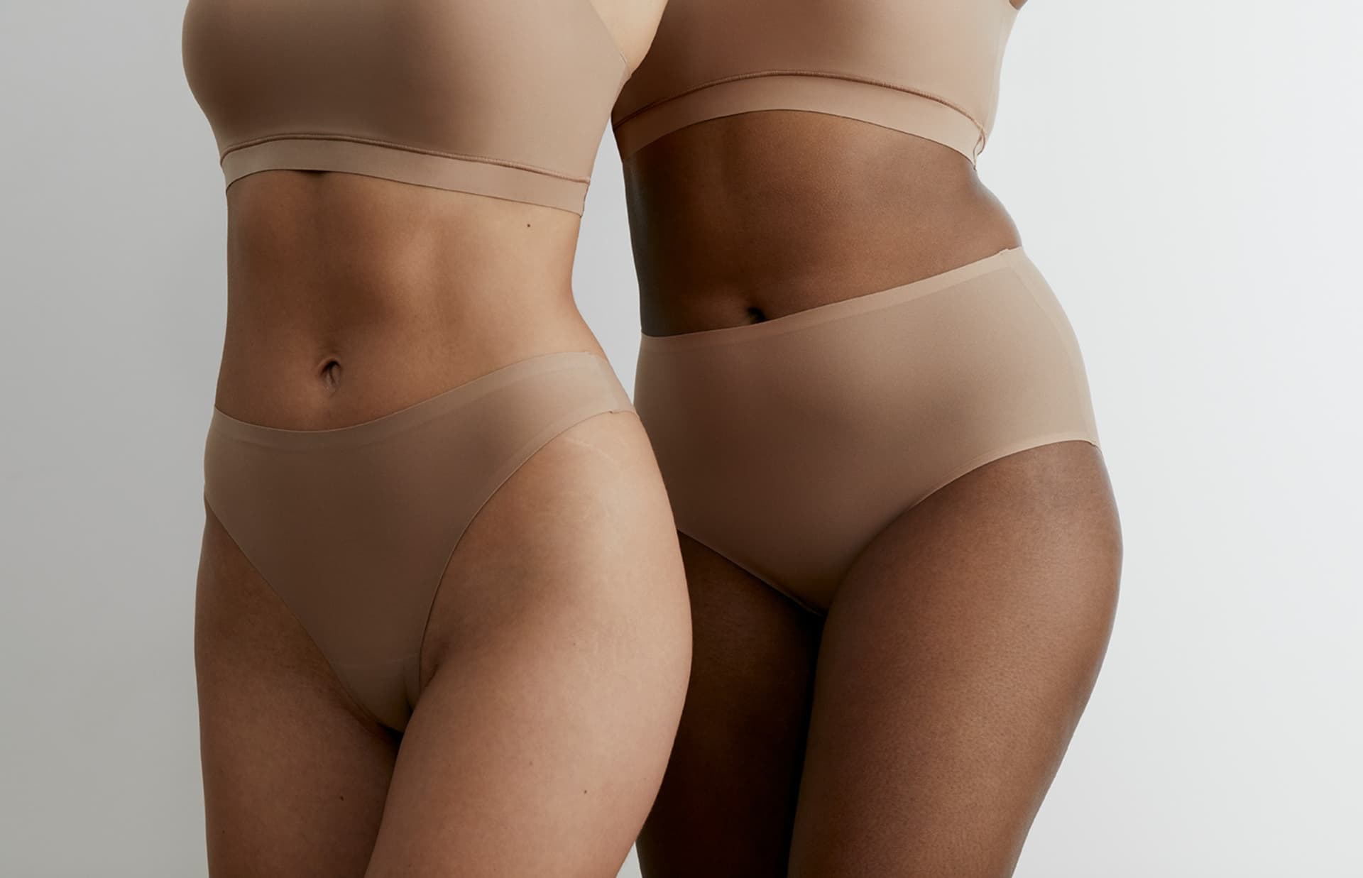 Two individuals in nude-toned underwear and bra are standing close together against a neutral background.