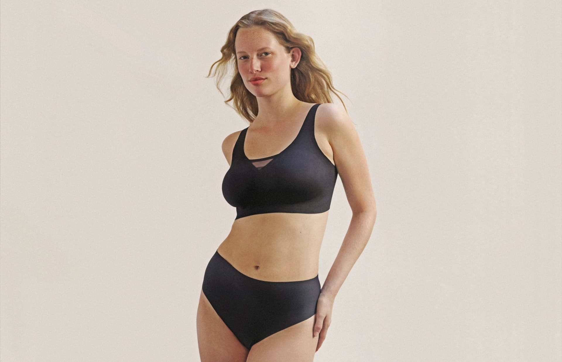 Woman in black underwear set, with long hair, standing against a plain background. Arms at her sides, confident expression.