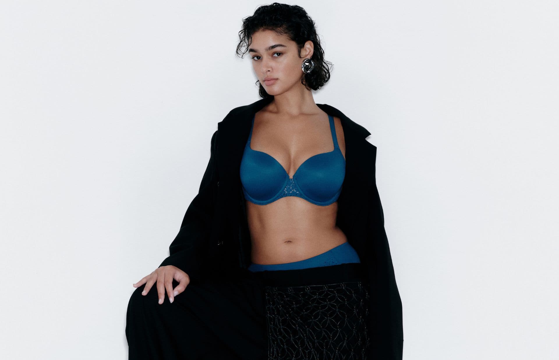 A person in a teal bra and underwear set, wearing a black coat, poses confidently against a plain white background.