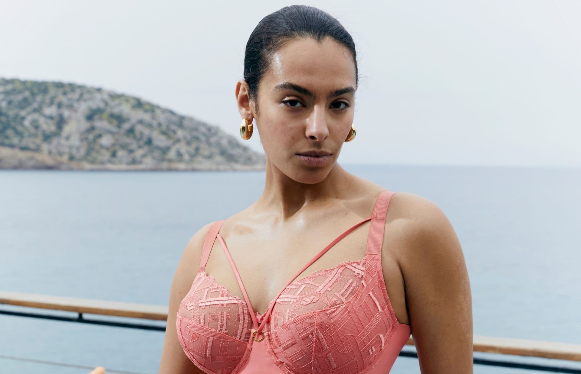 A person in a coral swimsuit stands on a deck with a scenic ocean and rocky hill in the background, wearing gold hoop earrings.