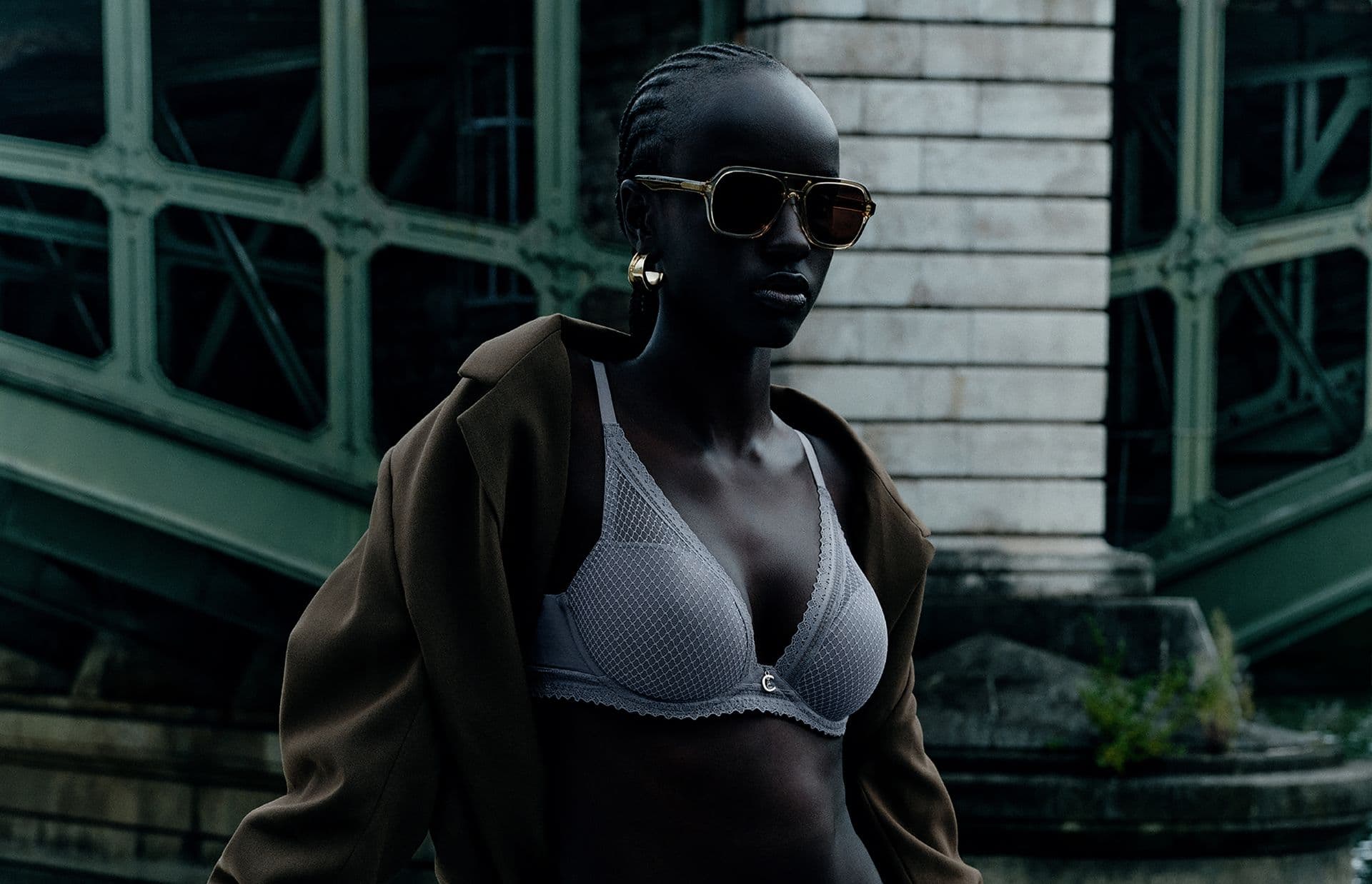 A person wearing sunglasses, a gray bra, and a coat poses confidently outdoors near a large, metal structure.