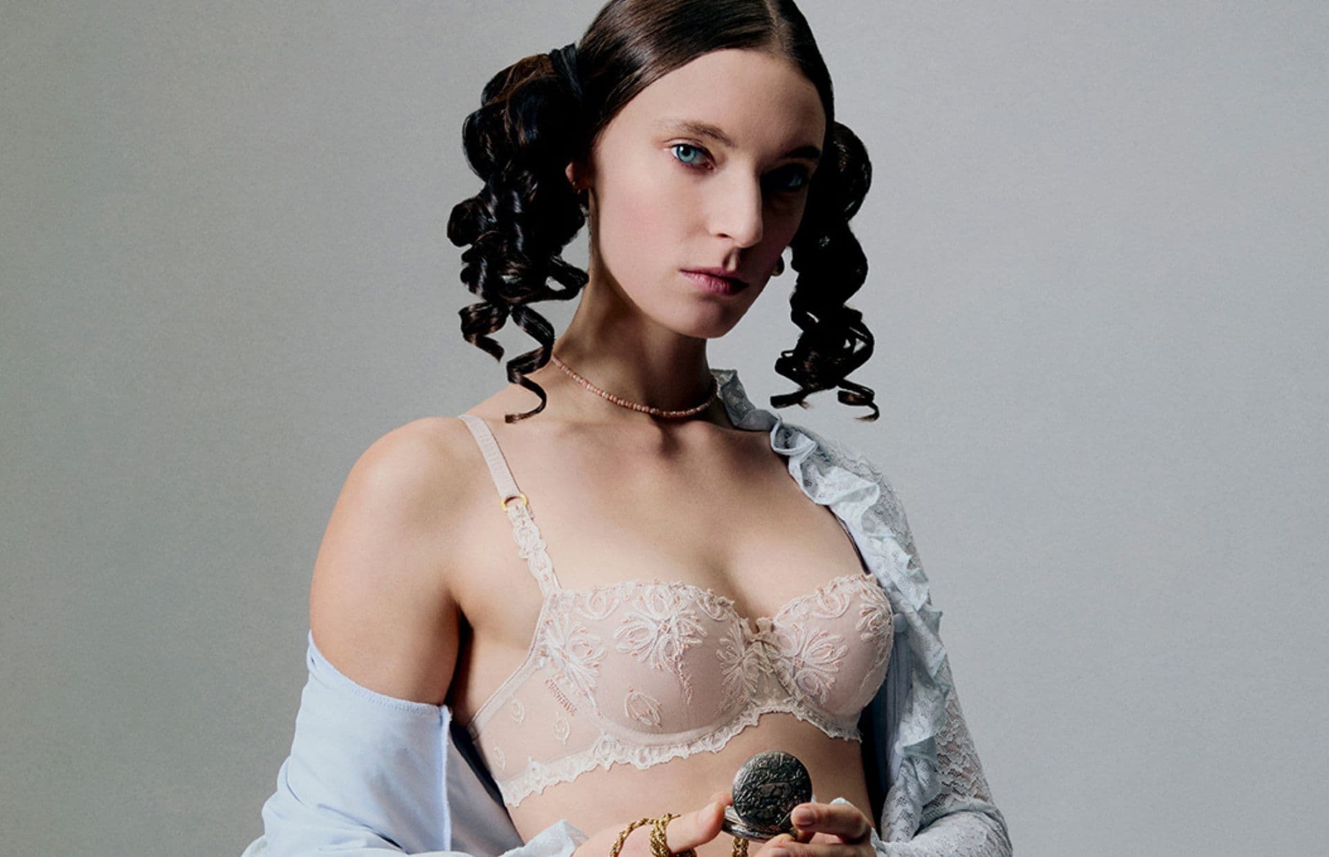 Person with curled hair wearing a lace bra and partially open shirt, holding an object, against a plain background.
