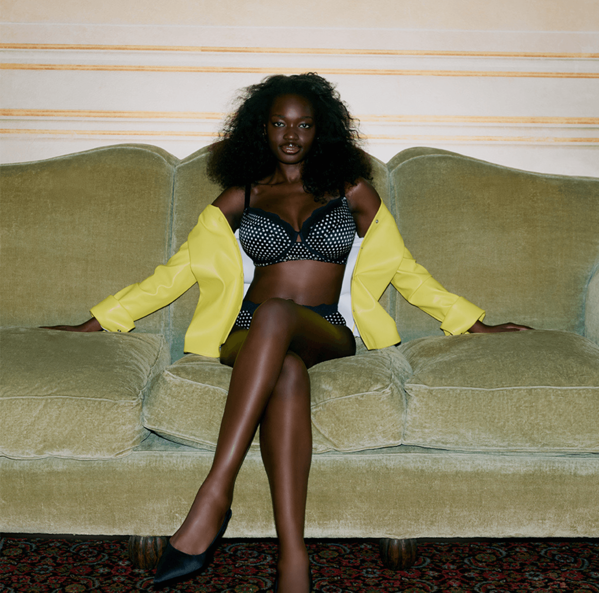 A woman in polka-dot lingerie and a yellow jacket sits confidently on a vintage green sofa.