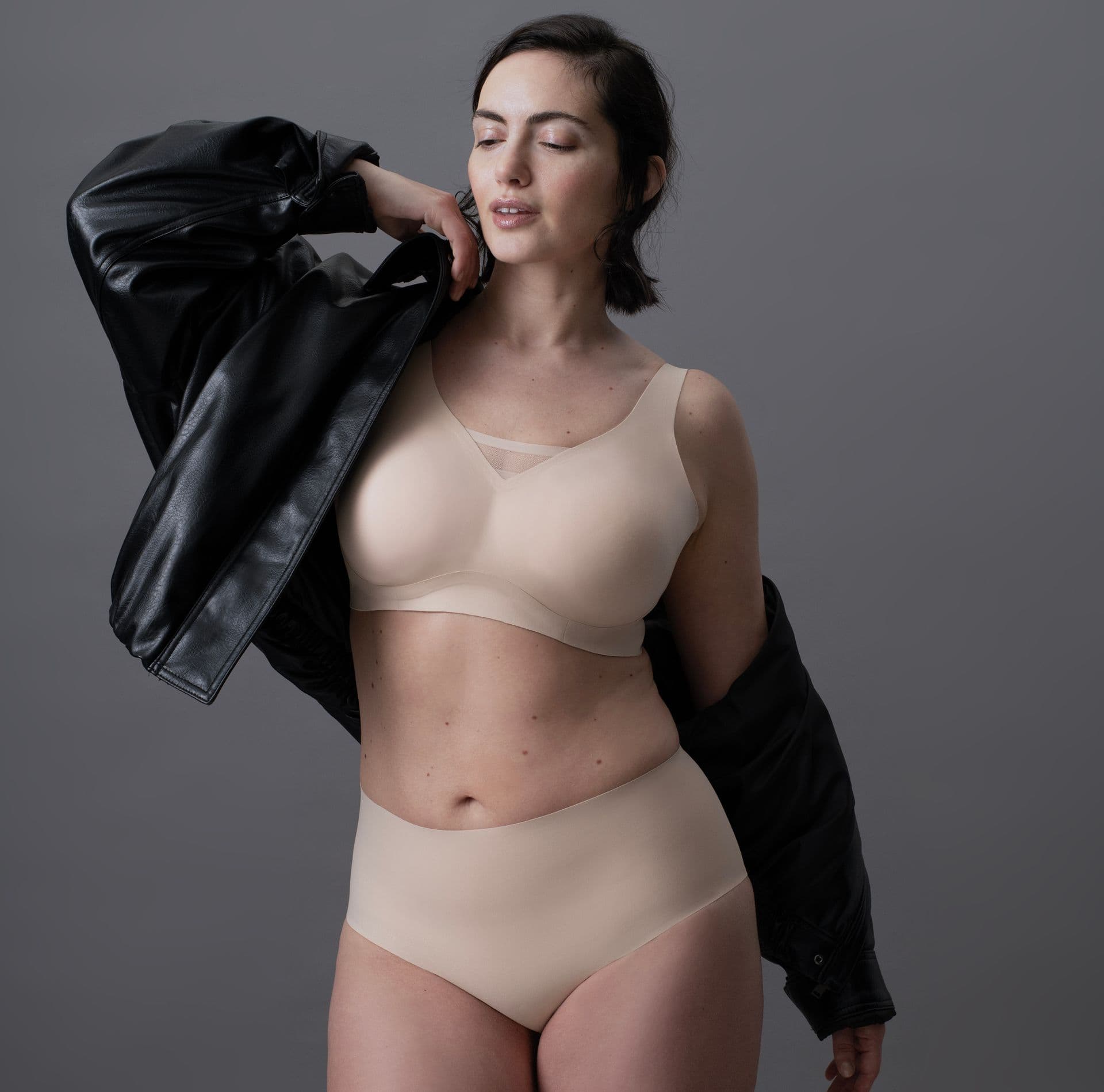 A woman in nude-colored lingerie and a black leather jacket poses confidently against a neutral gray background.