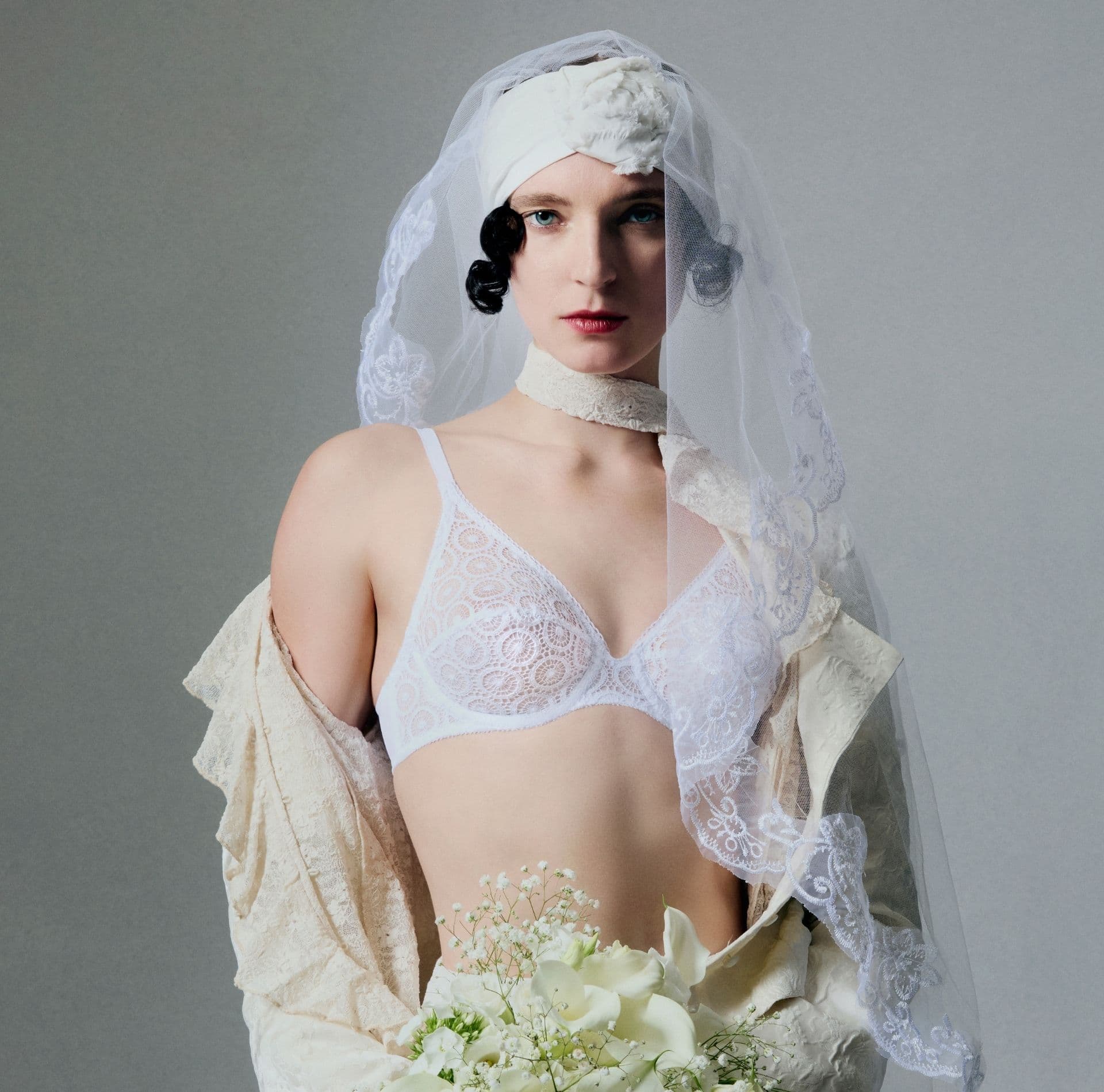 Person wearing a white bra, holding a bouquet of white flowers against a gray background.