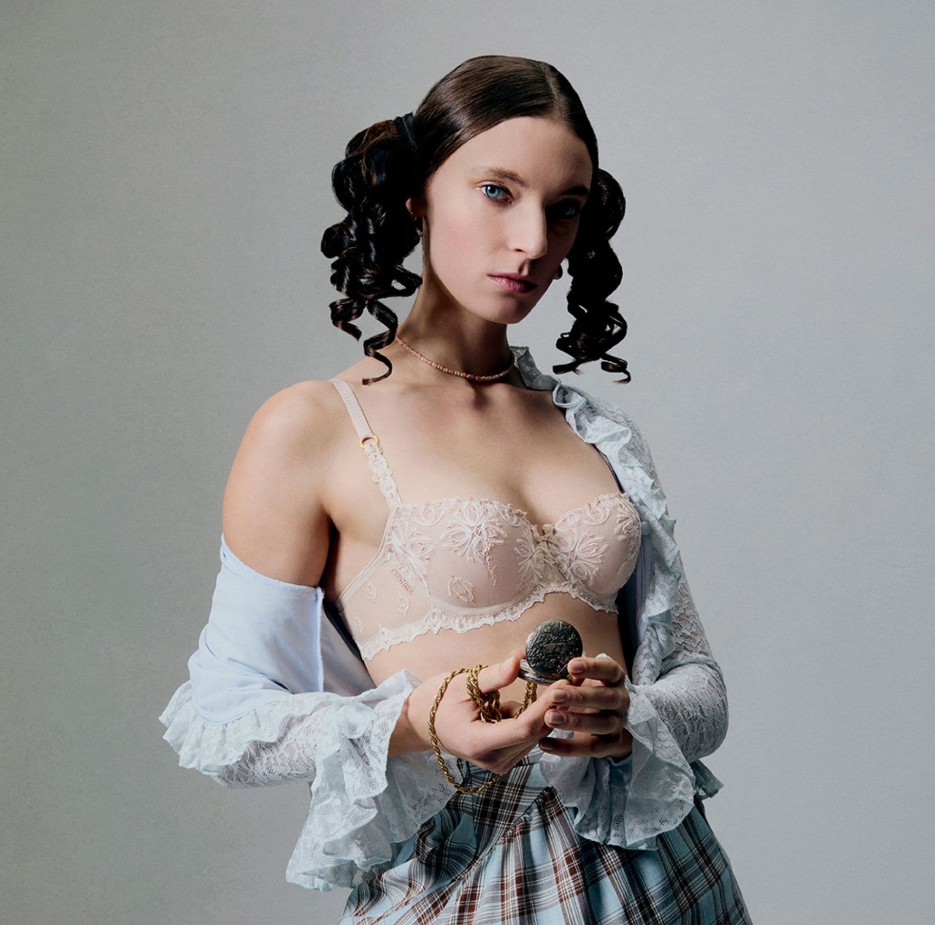 A woman with curled hair wears a lace top and plaid skirt, holding a small round object against a plain background.
