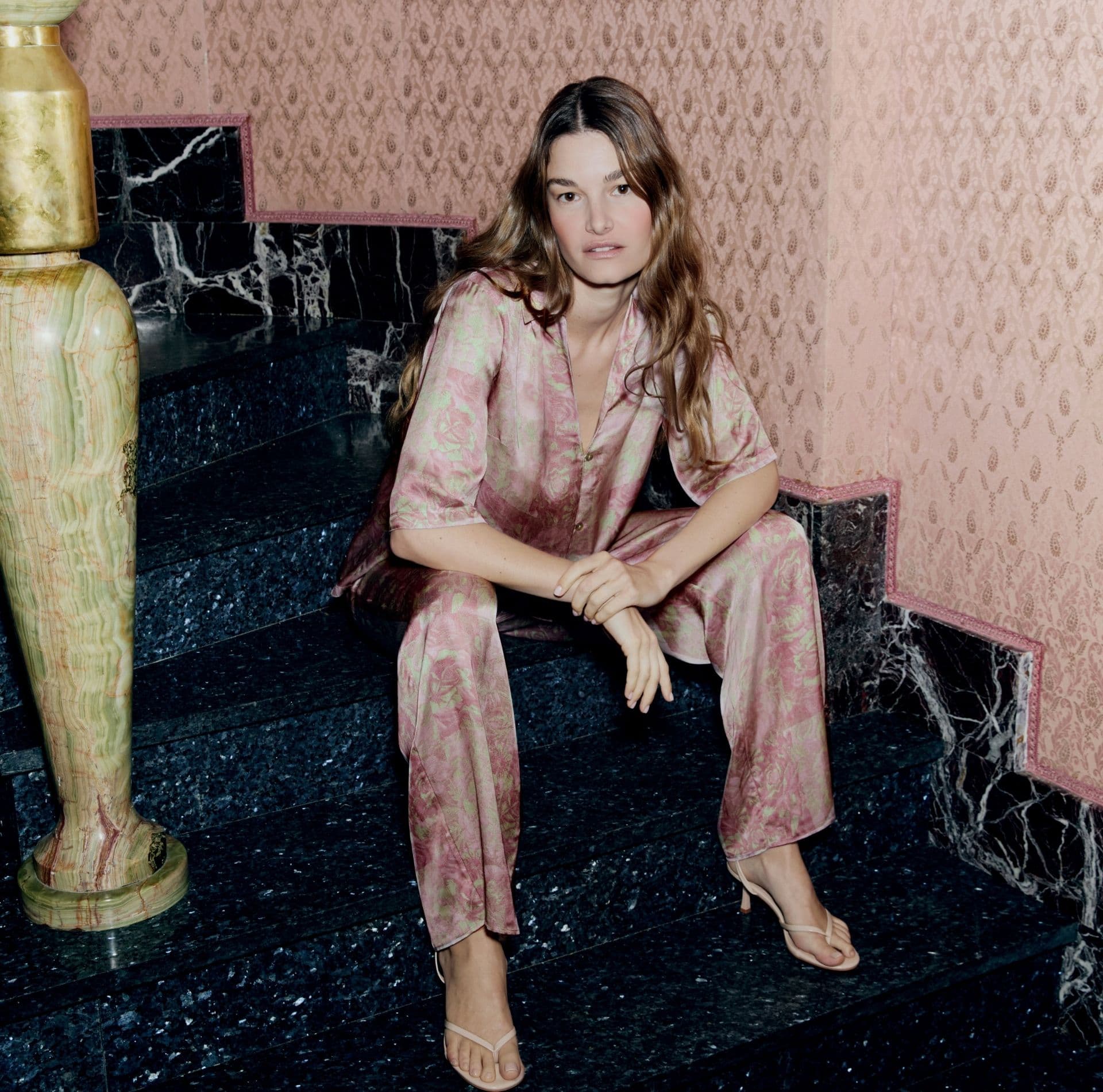 Woman sitting on black marble stairs, wearing a patterned pink and gold outfit, with long wavy hair, in an ornate interior setting.