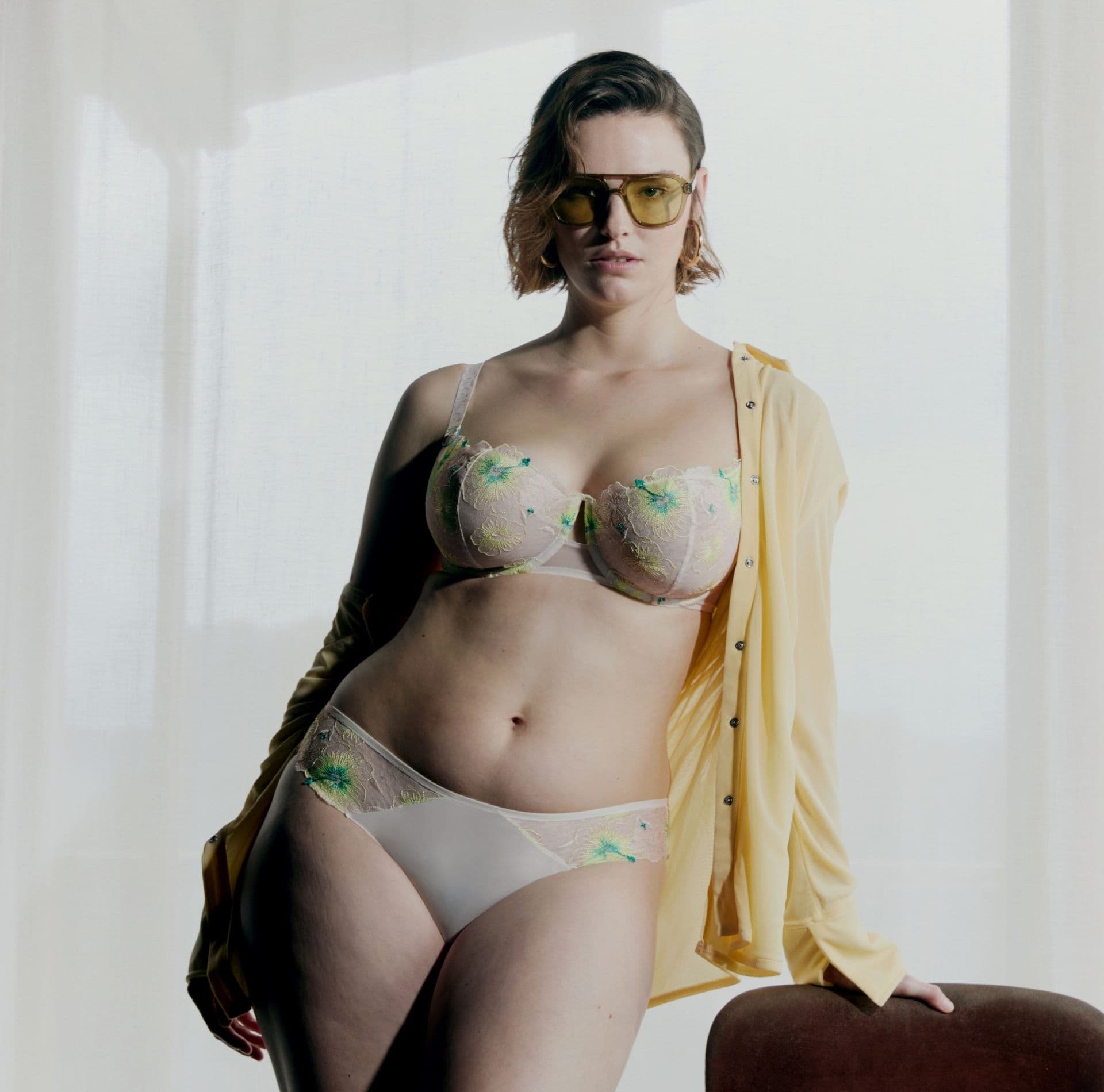 Person in floral lingerie and yellow shirt, wearing tinted glasses, poses confidently indoors with sheer curtains in the background.