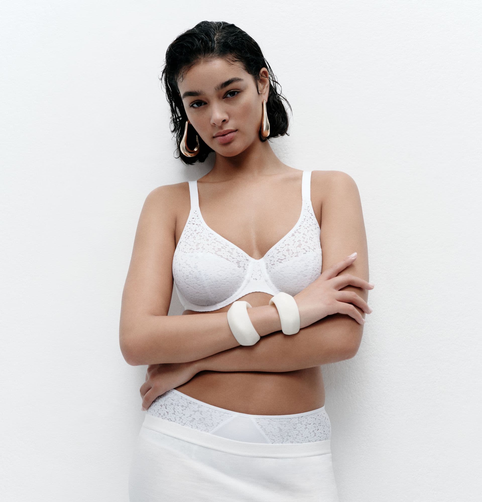 A woman wearing a white lace lingerie set stands with her arms crossed, styled with bold earrings and chunky bracelets against a minimal backdrop.