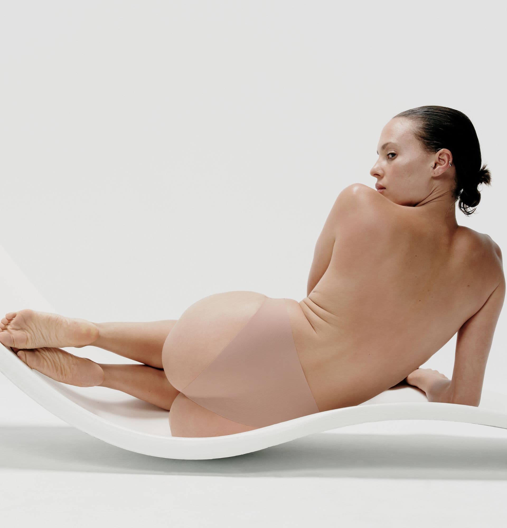 Person lying on a curved, white surface, wearing nude-colored underwear, against a plain white background.