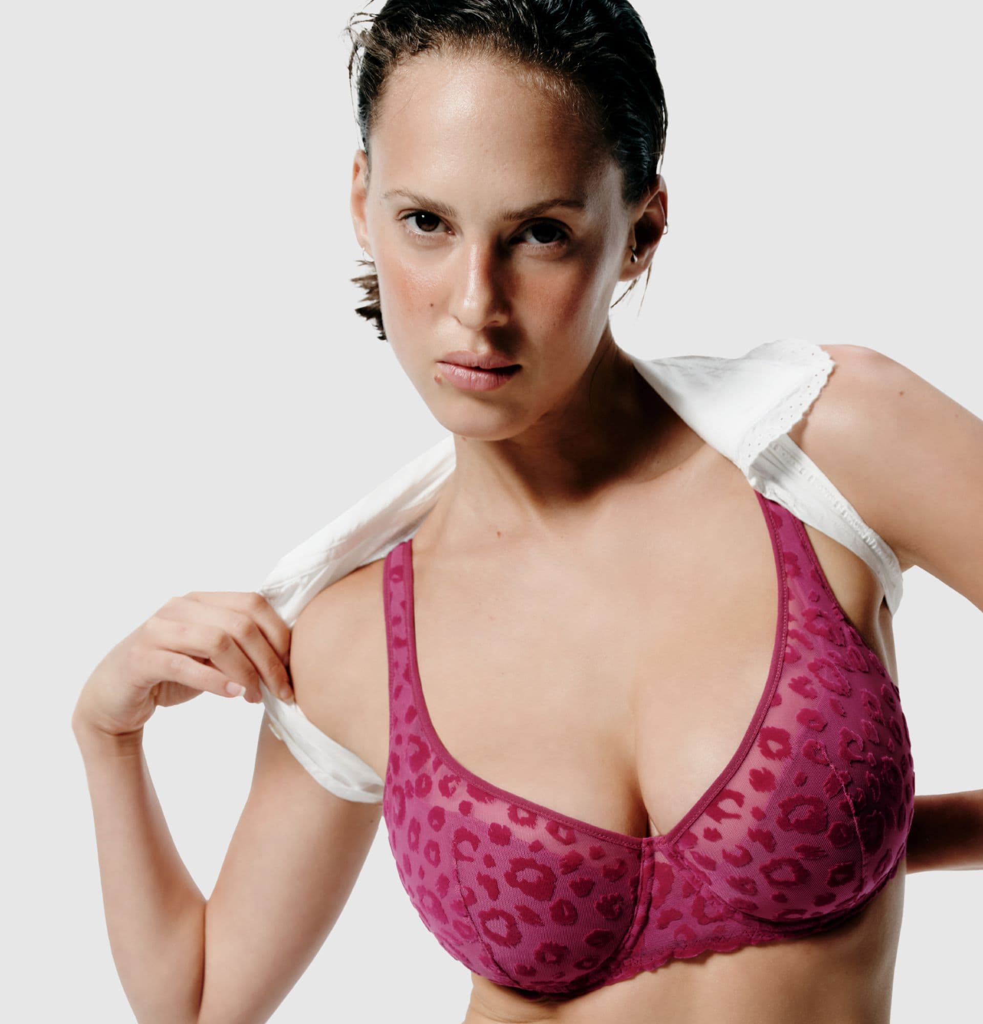 Person wearing a pink leopard-print bra, holding a white garment over their shoulder against a plain background.