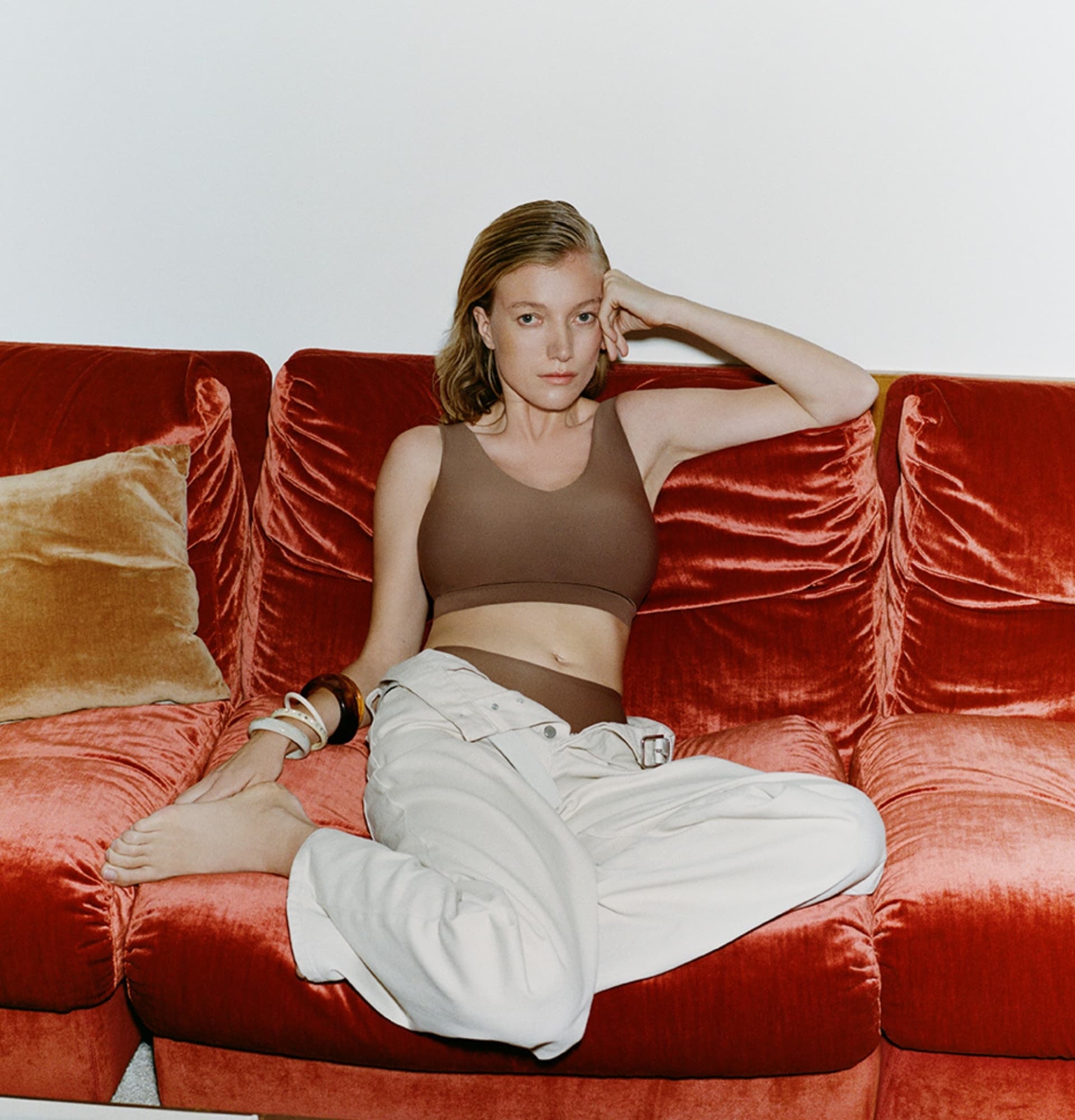 A person in a brown top and white pants lounges on a red sofa, with one hand on their head and wearing several bangles.