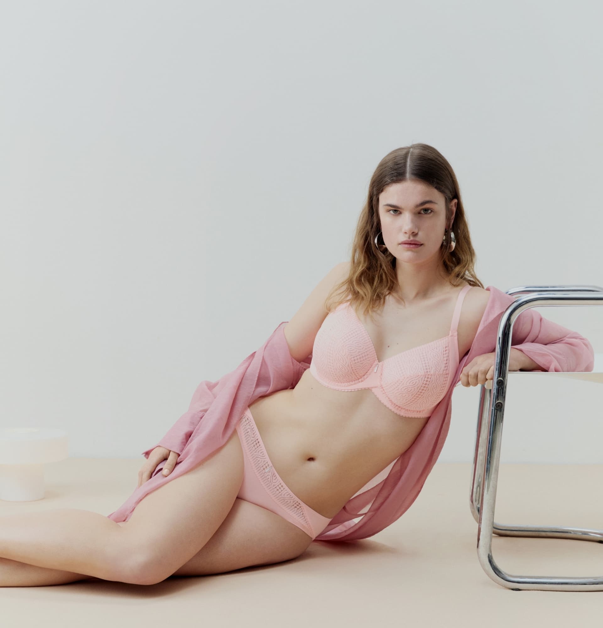 A woman in pink lingerie and an open shirt lounges on the floor next to a metal chair, set against a minimalist backdrop.