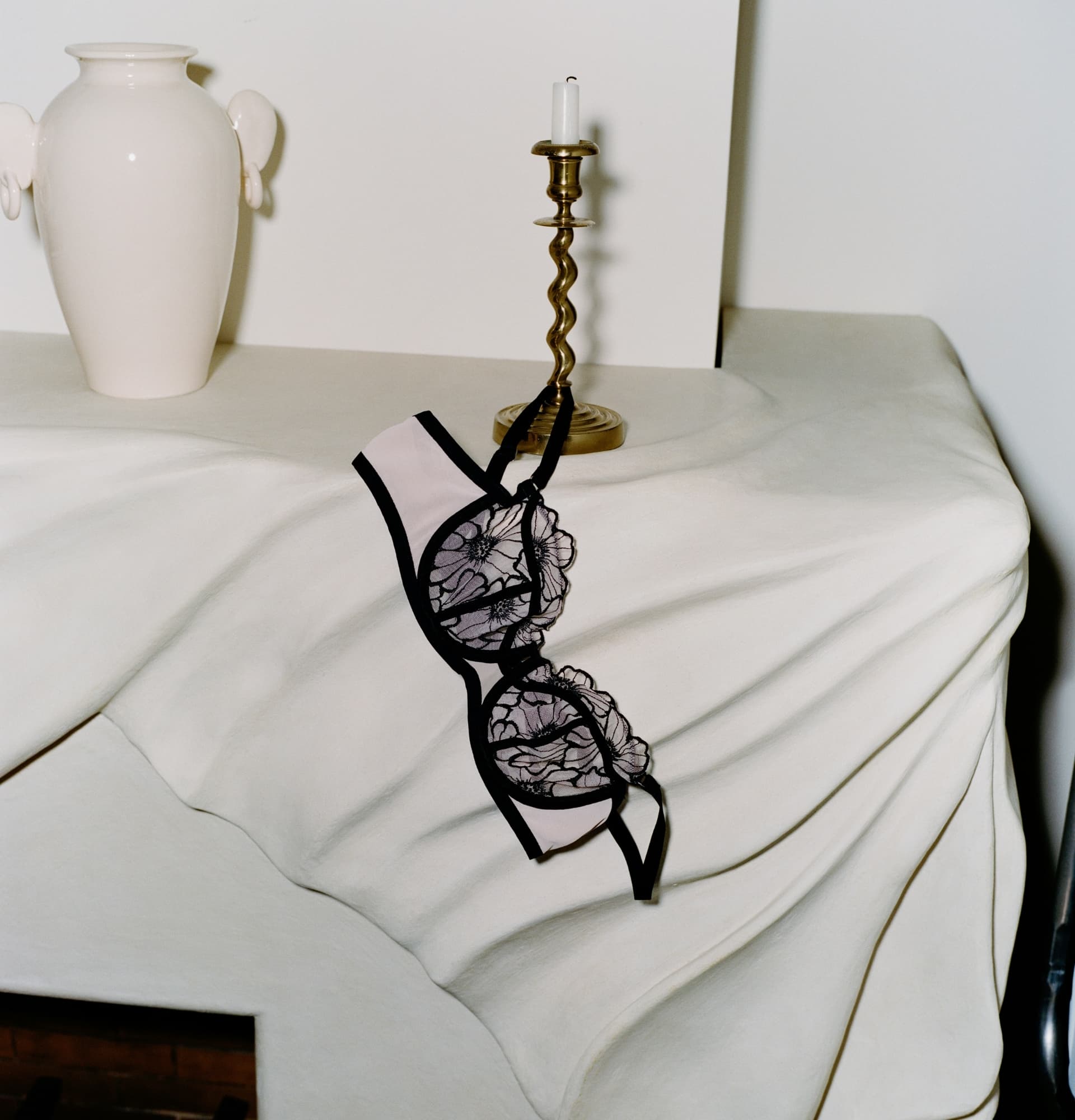 A lace bra with floral patterns draped over a white sculpted surface, beside a white vase and a brass candlestick with an unlit candle.