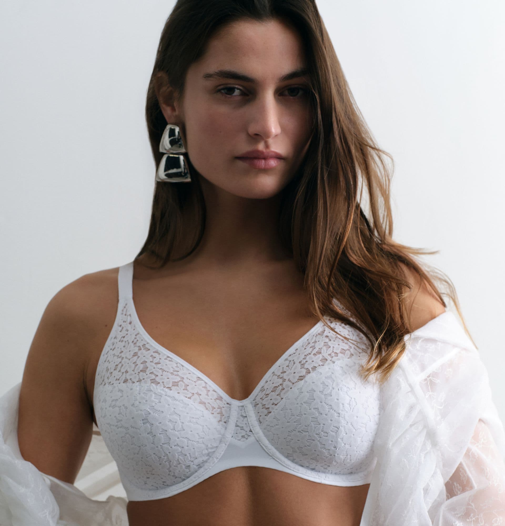 Woman wearing a white lace bra and large silver earrings, draped with a sheer white fabric, against a plain background.