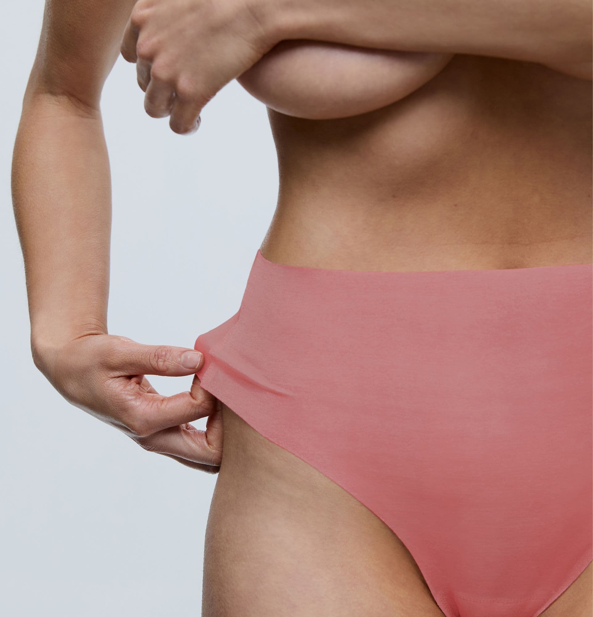 Person wearing coral high-waisted underwear, adjusting the waistband with one hand, against a light background.