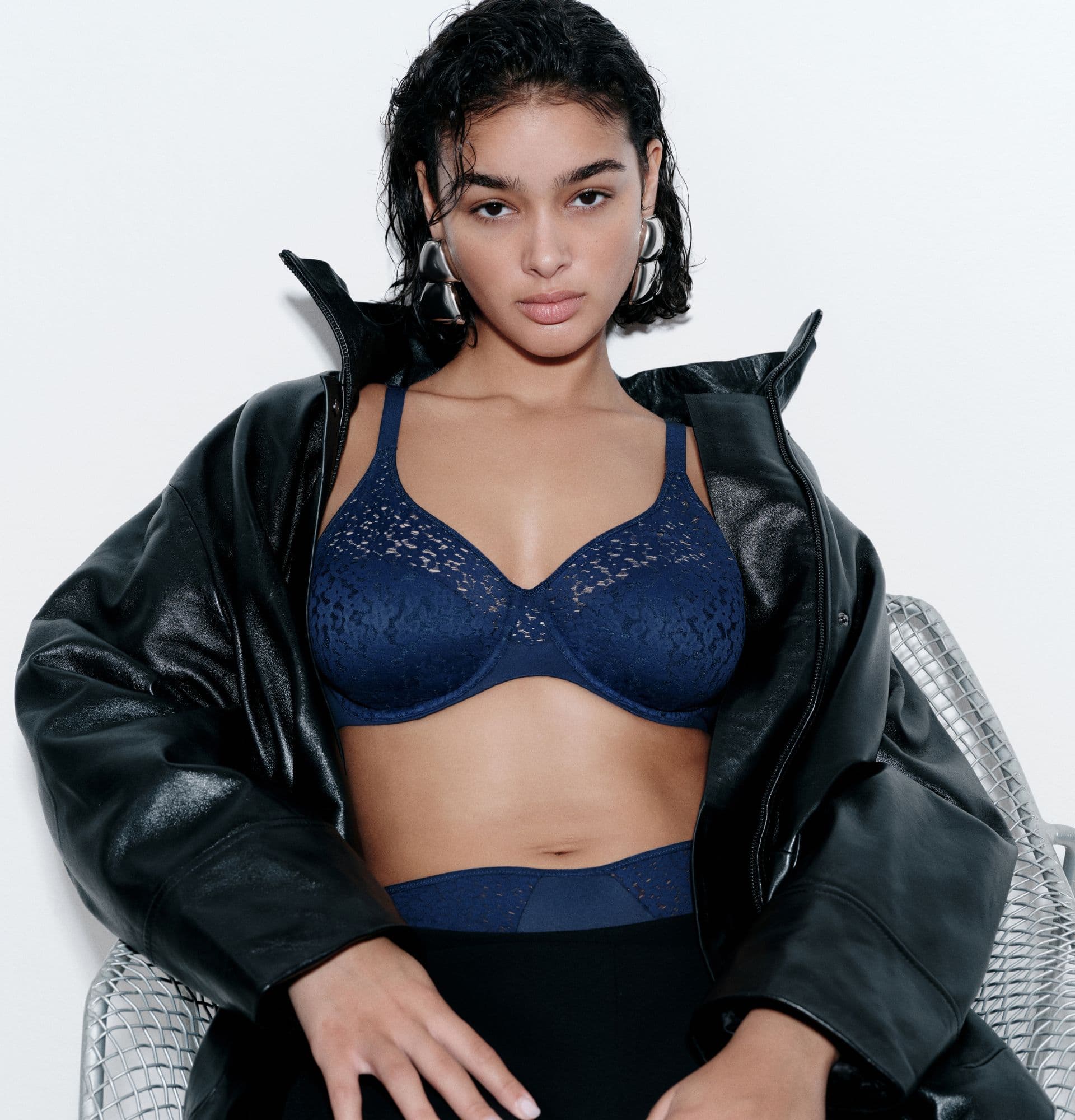 Person wearing a navy blue lace bra and black leather jacket, sitting on a silver chair against a white background.
