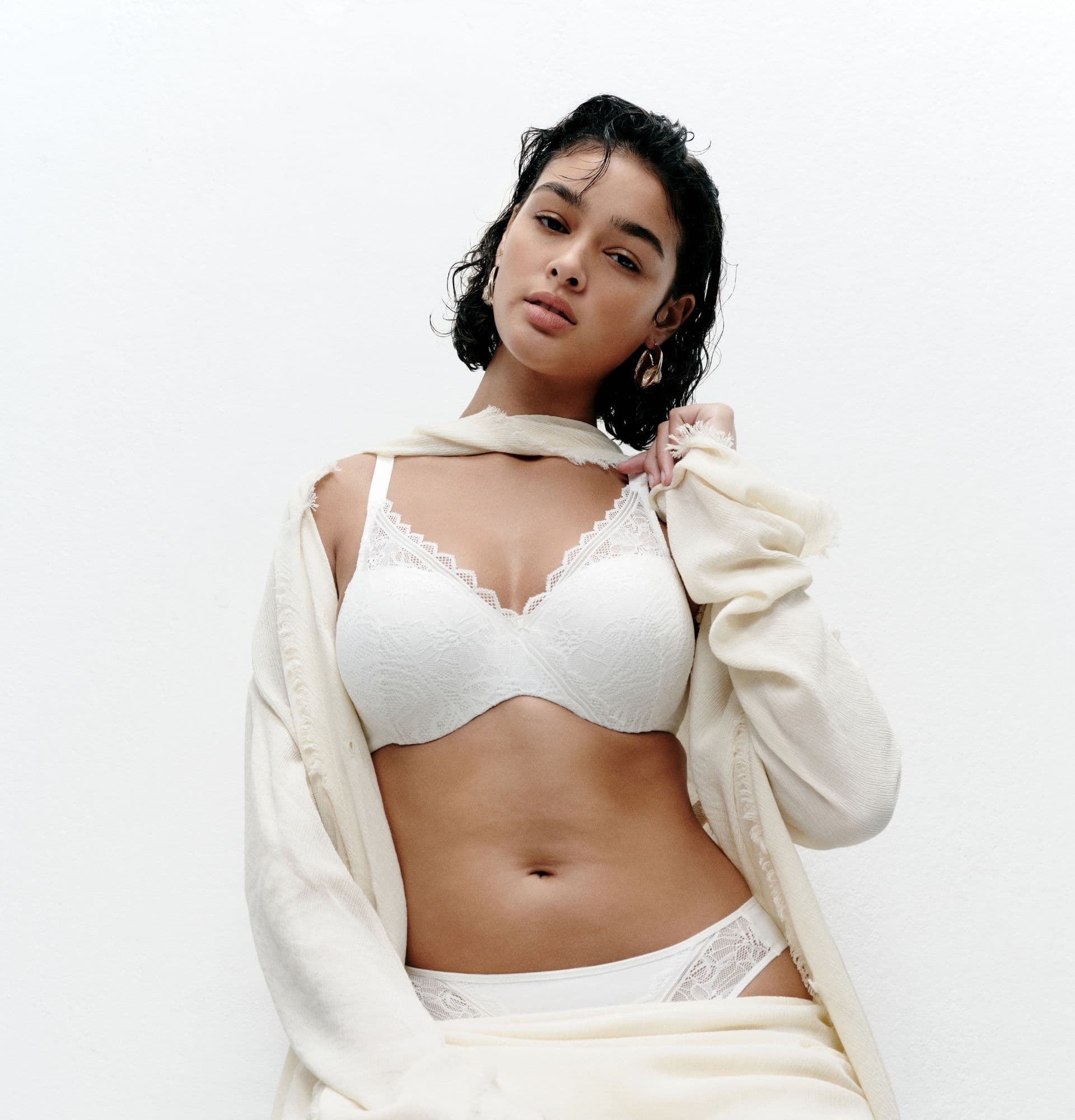 Woman in white lace lingerie and a cream shawl poses confidently against a plain white background.