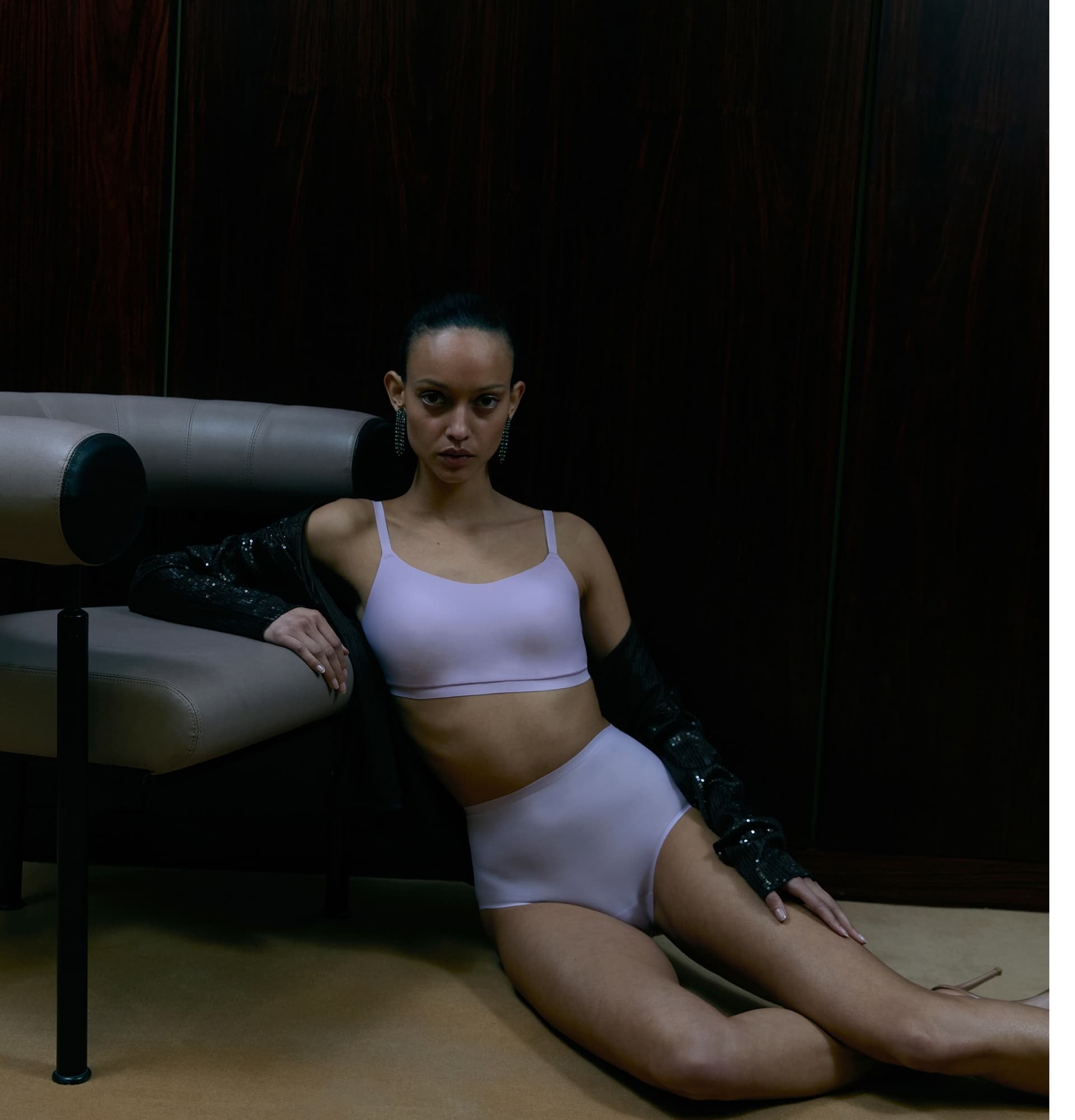 Person in light purple lingerie sits on the floor, leaning against a modern chair, with a dark jacket draped over their shoulders.