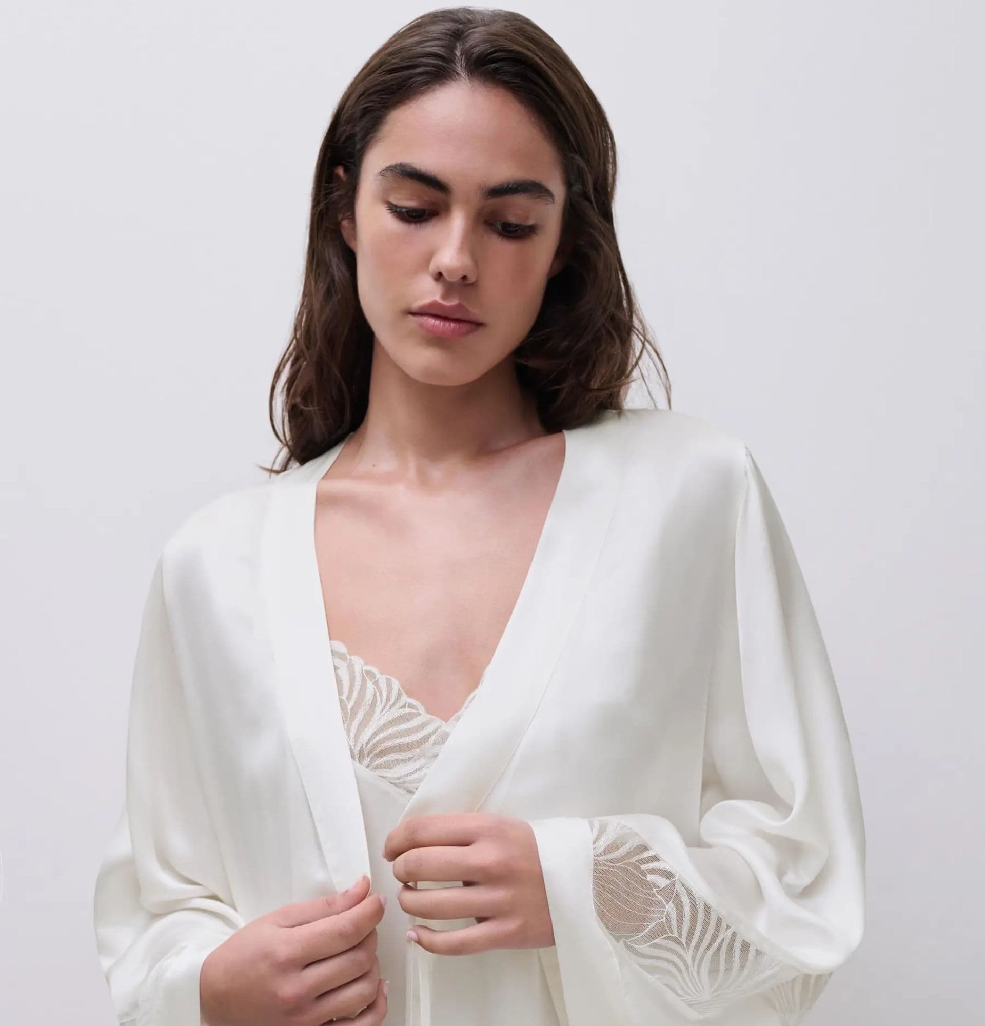 Woman in a white robe with delicate lace details, looking downwards against a plain background.