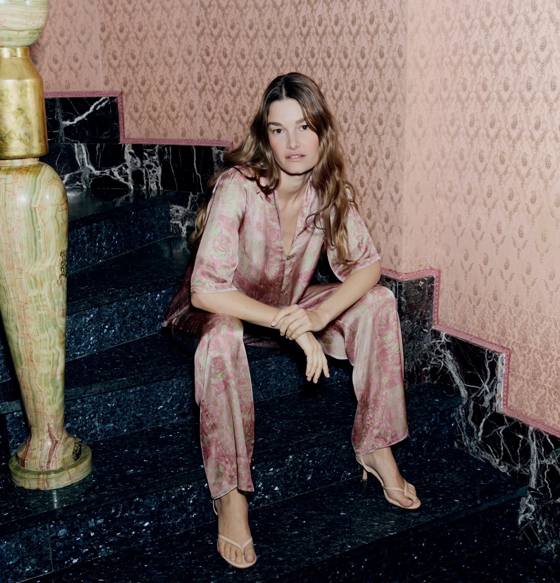 Woman in a pink patterned outfit sits on a marble staircase with ornate wallpaper in the background.