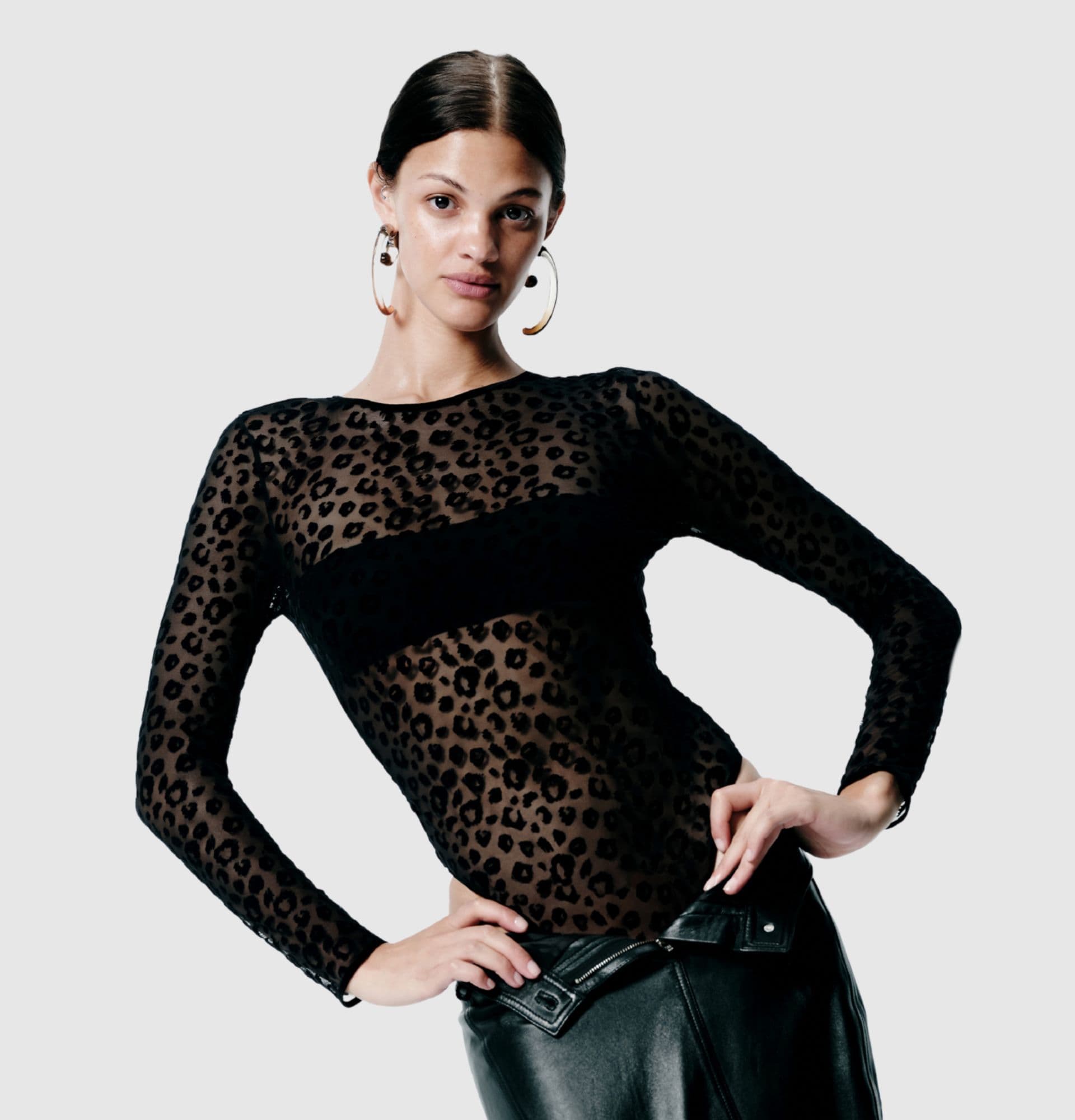 Person wearing a sheer leopard-print top and large hoop earrings, posing with hands on hips against a plain background.