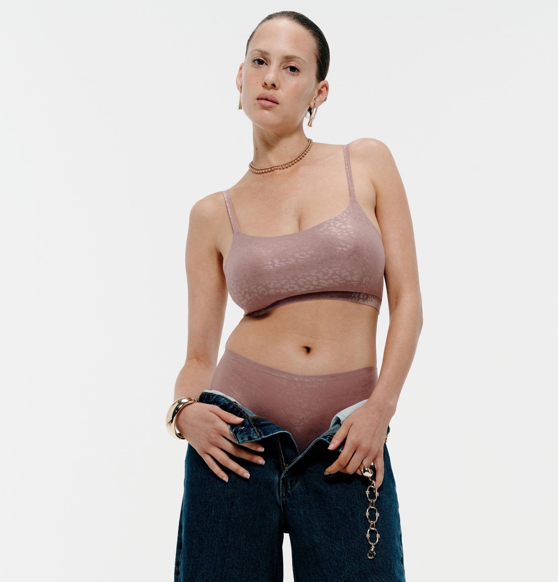 Person in a mauve undergarment set, wearing gold earrings and bracelets, holds open unbuttoned jeans against a white background.