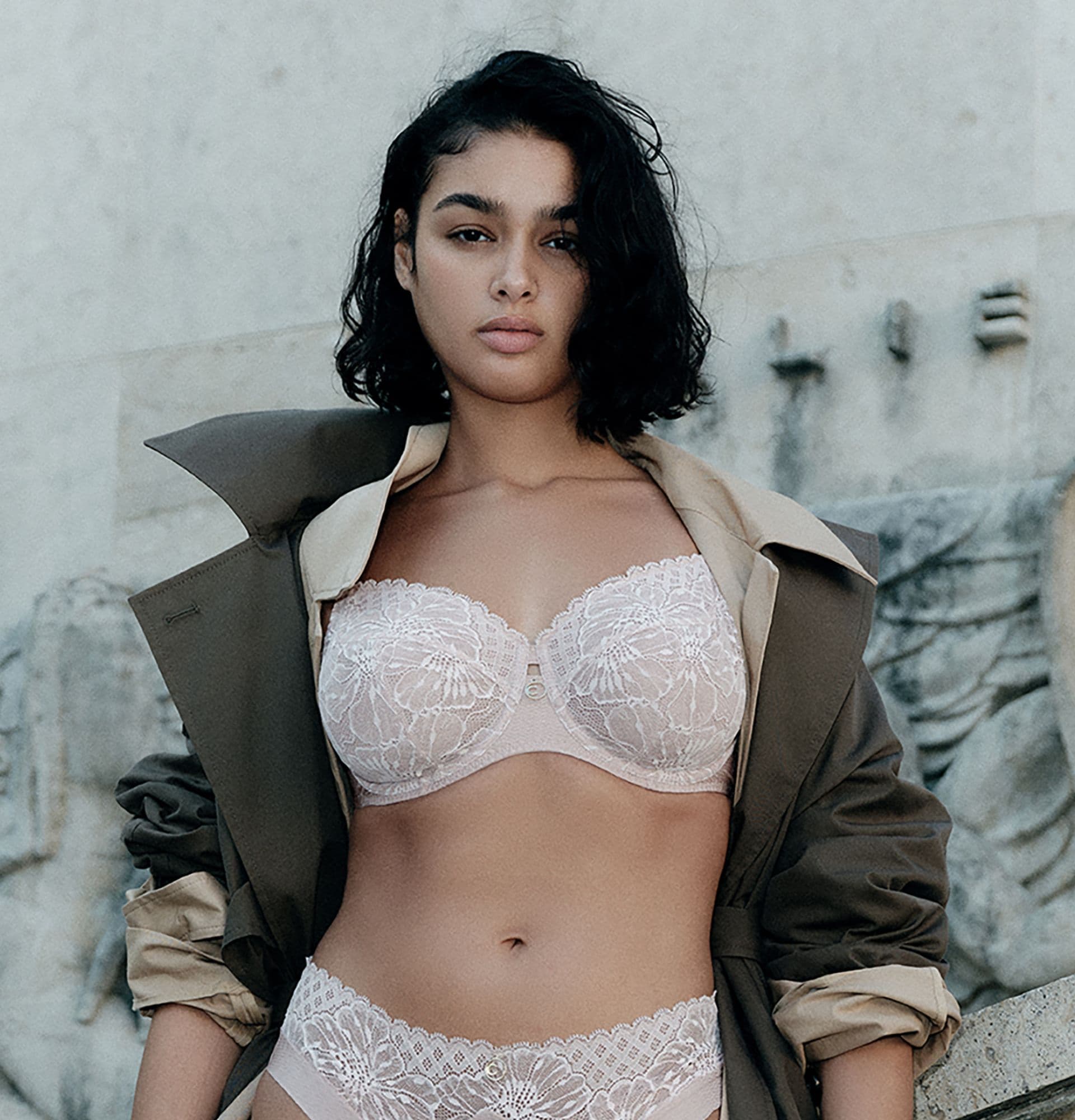 A woman wearing a lace lingerie set and an open coat stands confidently outdoors against a stone wall backdrop.