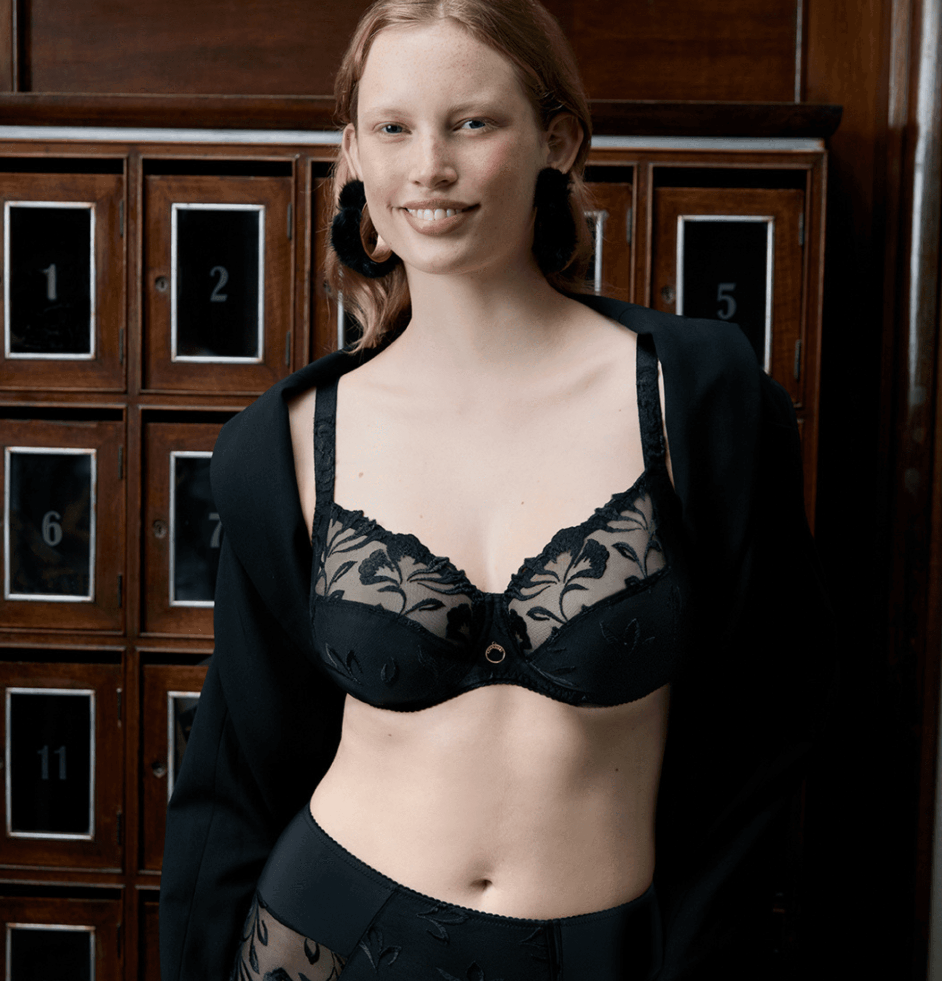 A person wearing black floral lace lingerie and an open jacket stands in front of a wall with numbered boxes.