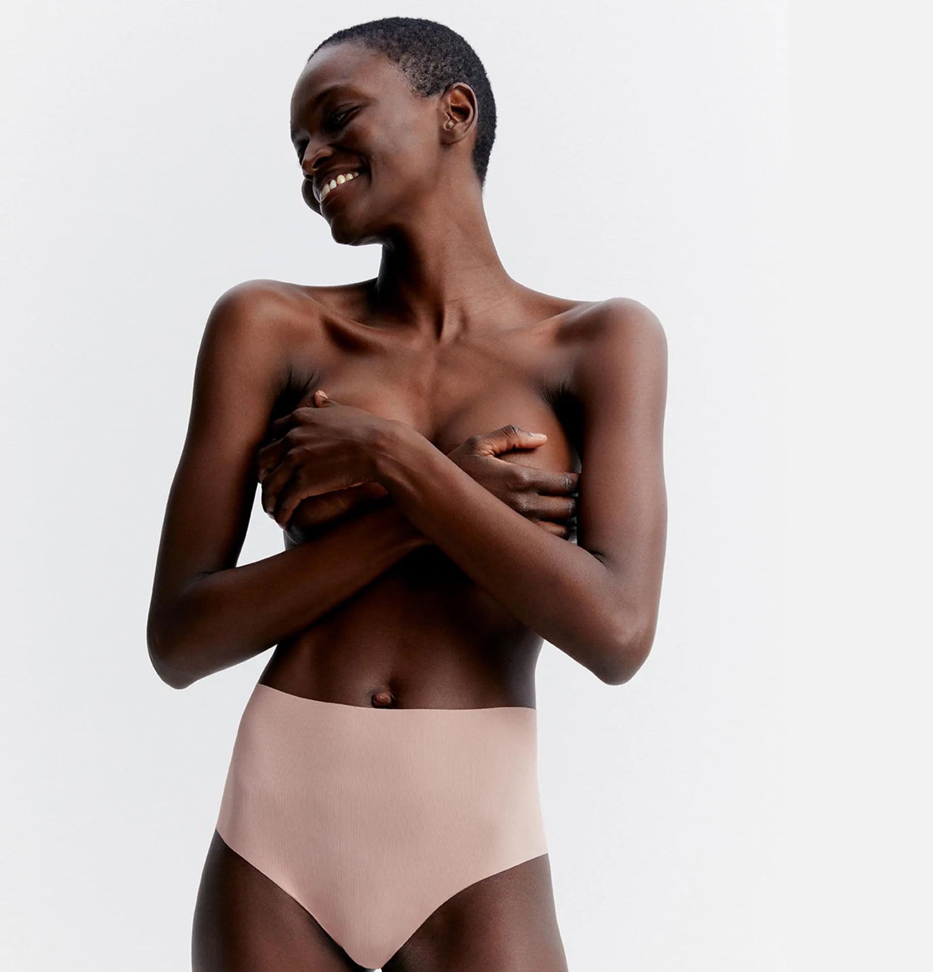 Smiling person with short hair covers chest with arms, wearing high-waisted nude undergarment, standing against a light background.