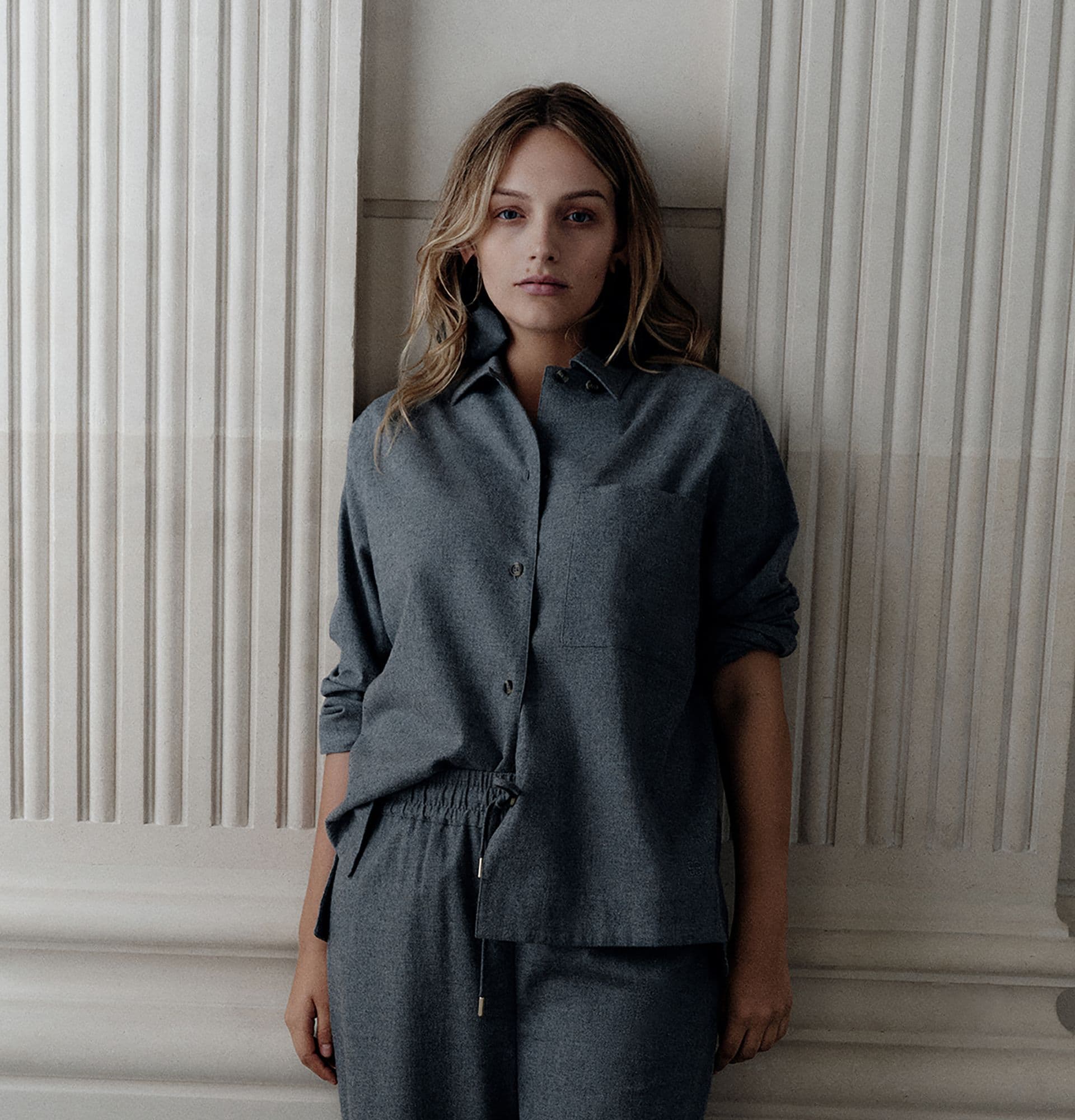 Woman in a loose-fitting gray outfit stands against a textured wall, gazing forward with a neutral expression.