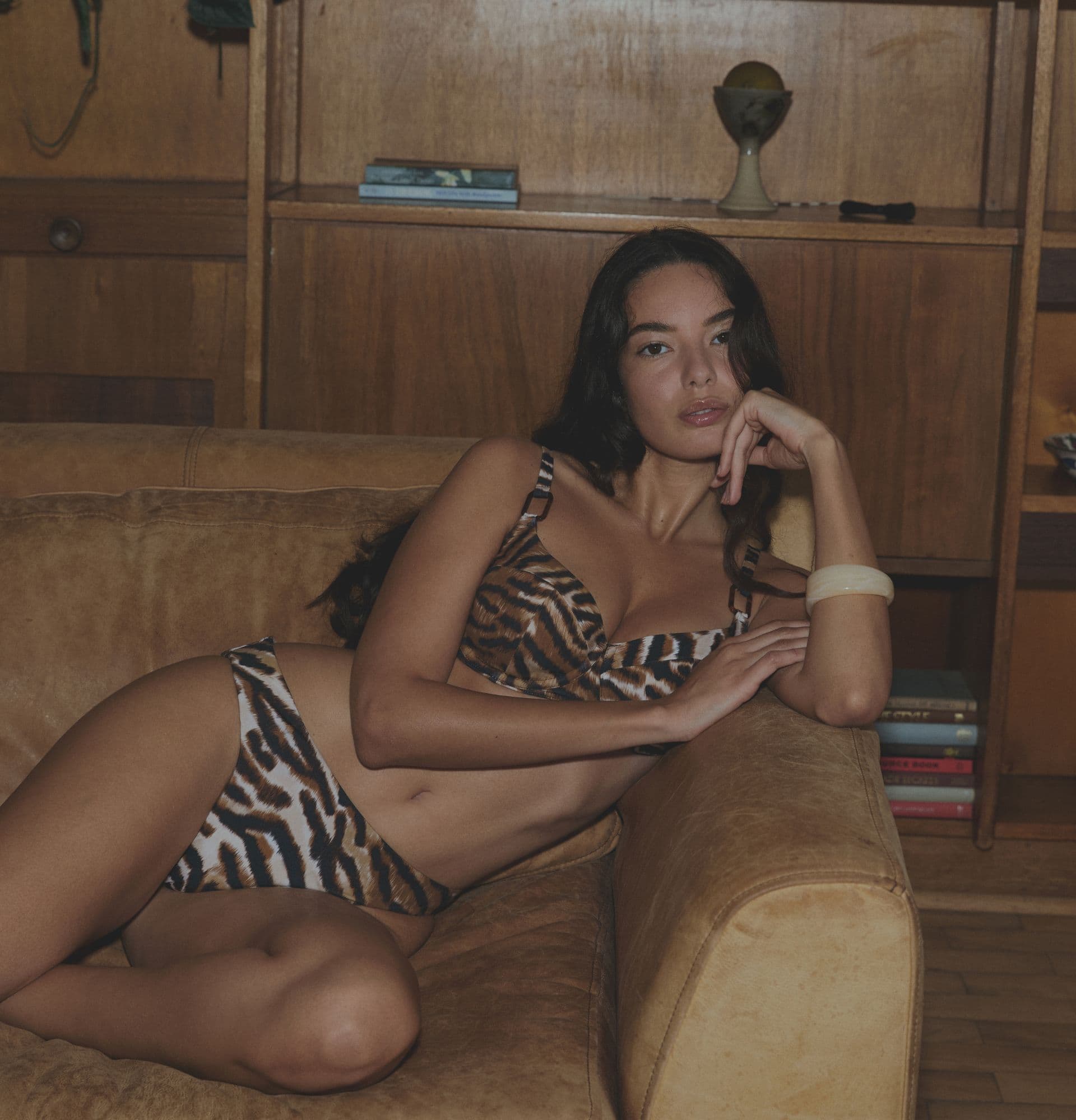 Person lounging on a tan couch wearing a tiger-print bikini, with a wooden shelf in the background.