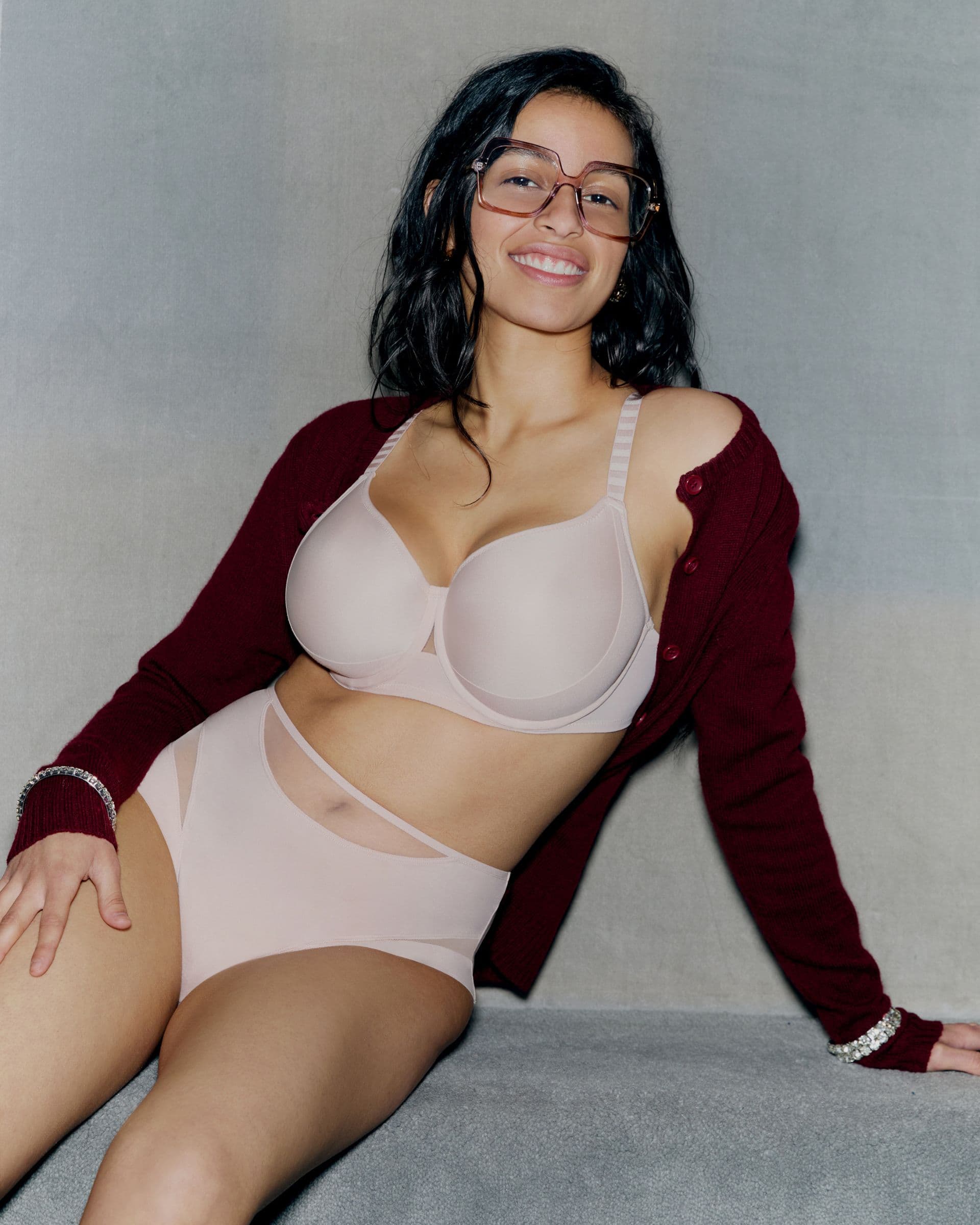 Person smiling, wearing glasses, a burgundy cardigan, and nude lingerie, sitting against a gray backdrop.
