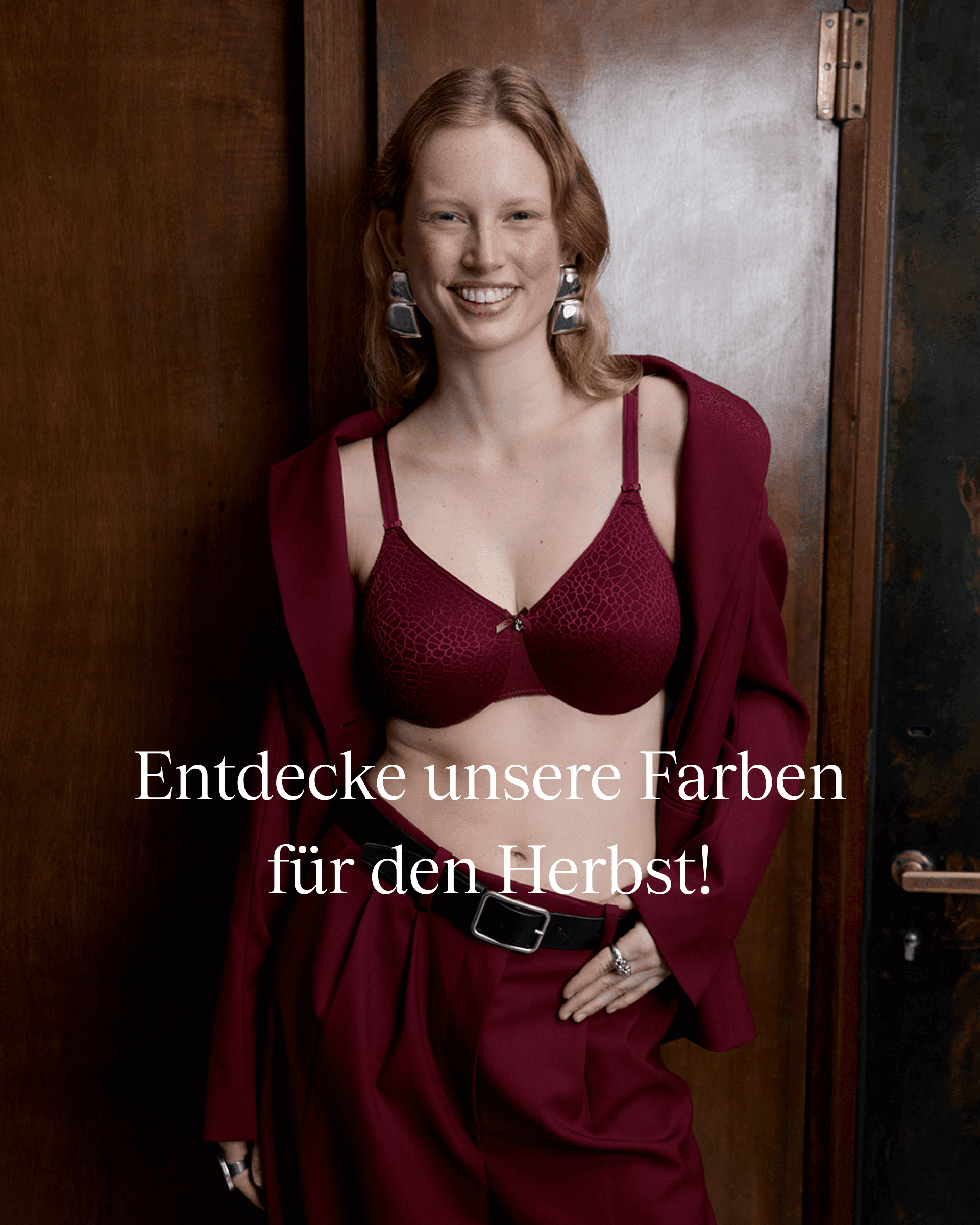 Smiling woman in a burgundy outfit stands by a wooden door, showcasing a stylish bra. Text promotes autumn colors in German.