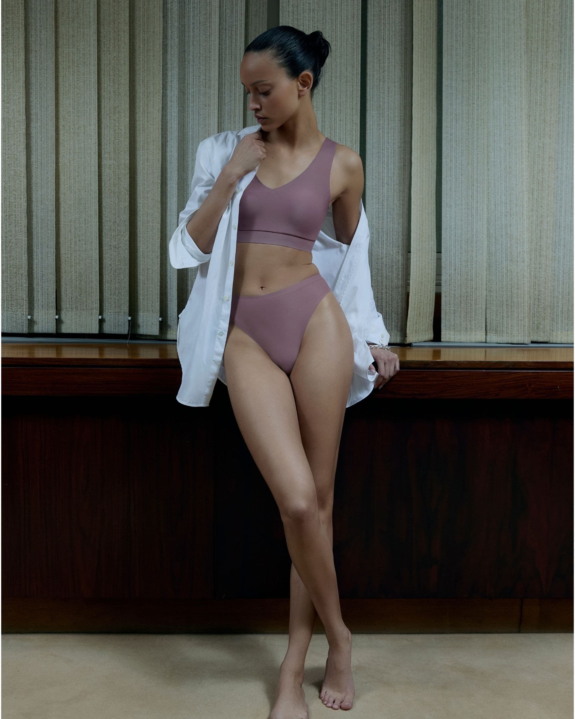A person in mauve underwear and a white shirt stands indoors, looking down, against a background of vertical blinds.