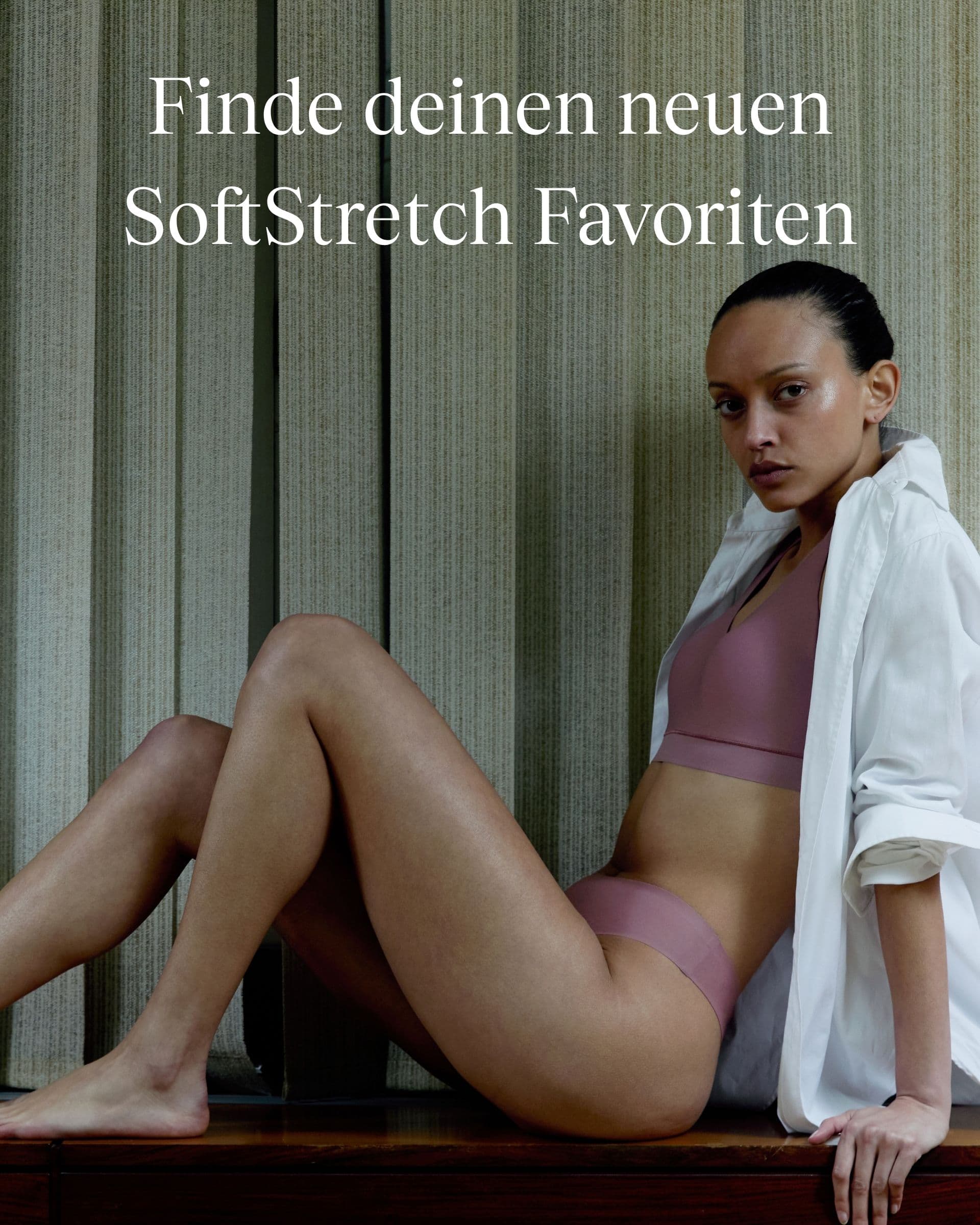 Person in a mauve undergarment set and white shirt sits on a wooden bench in front of vertical blinds. Text reads "Finde deinen neuen SoftStretch Favoriten".