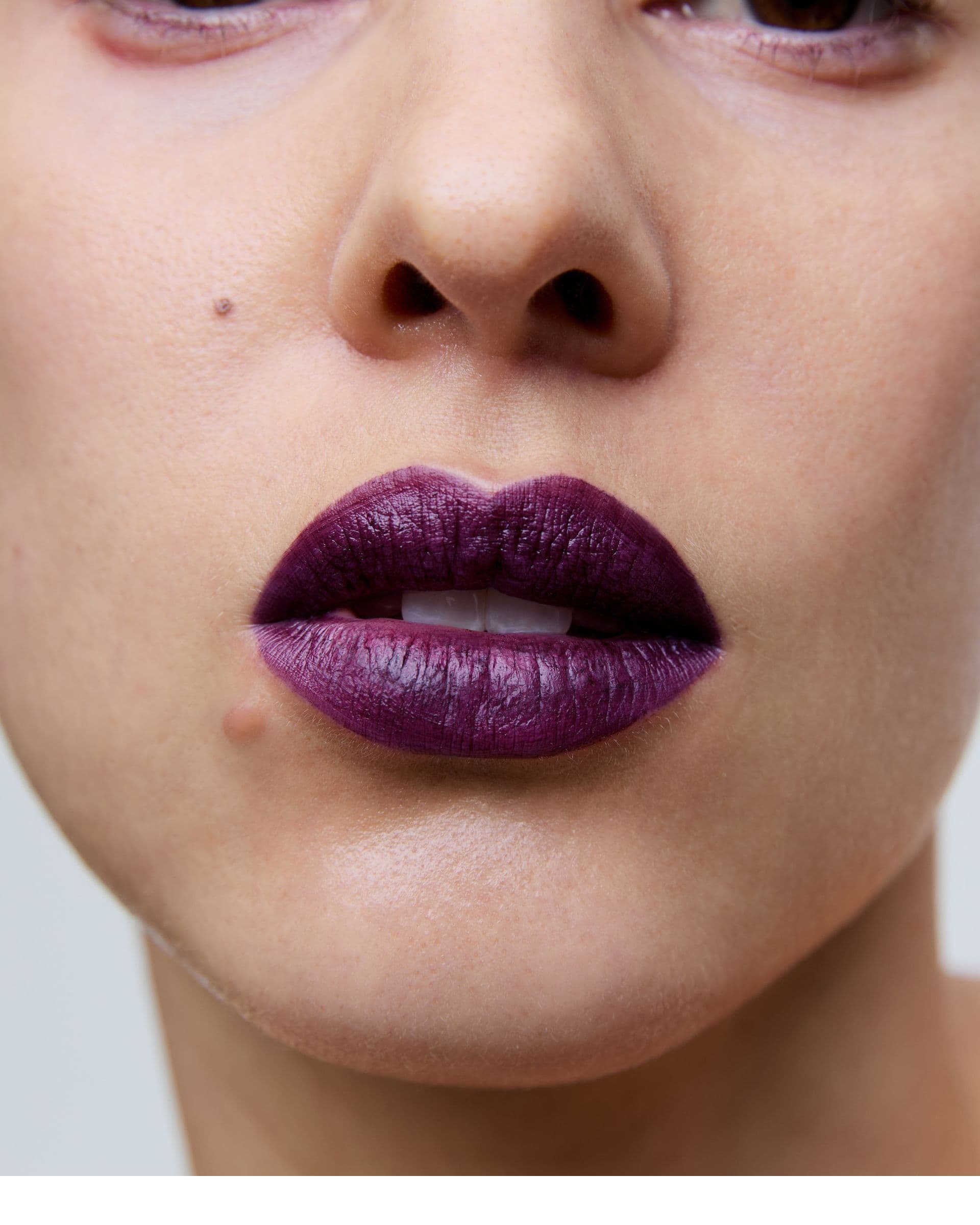 Close-up of a person's lips with dark purple lipstick, showcasing a small beauty mark and natural skin texture.