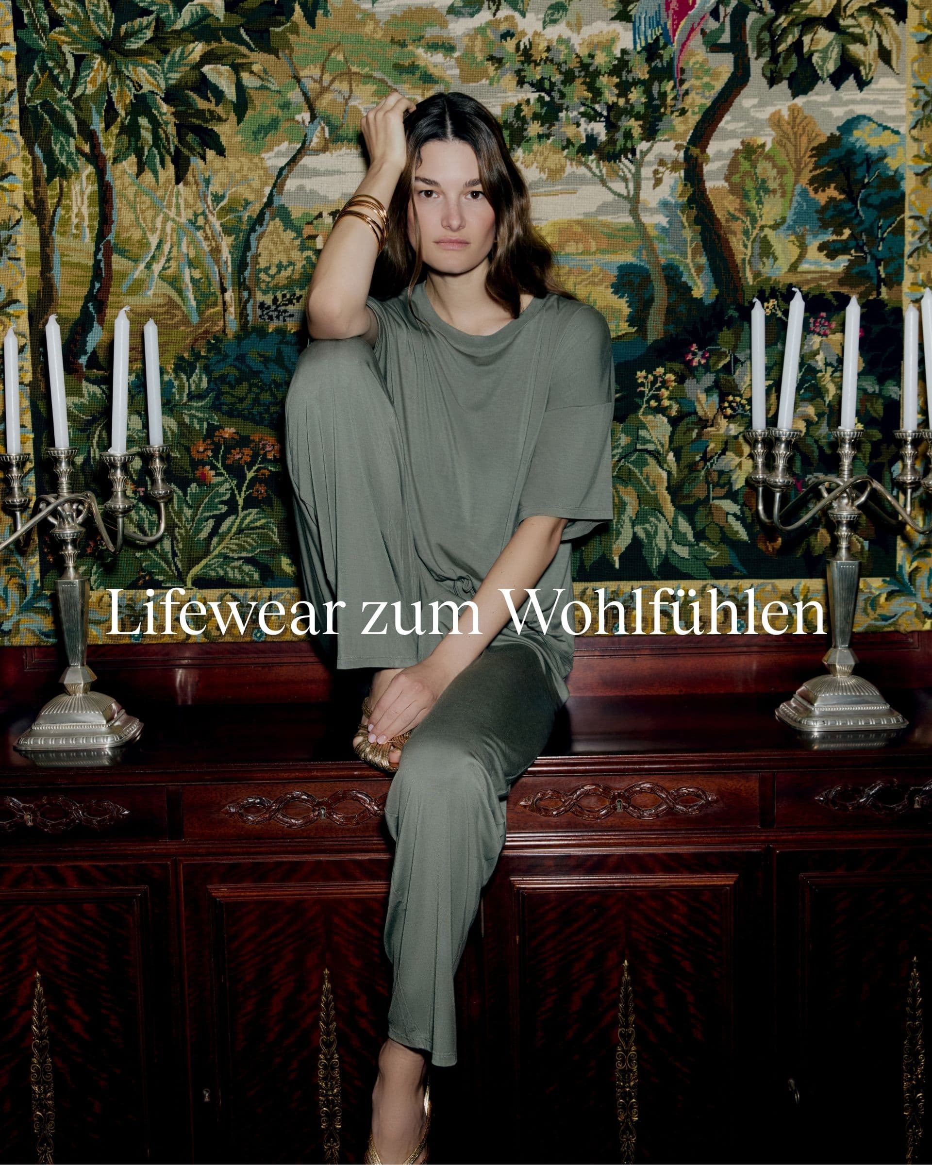 Woman in a green outfit sitting on a wooden surface with ornate wallpaper and candelabras, text reads "Lifewear zum Wohlfühlen."
