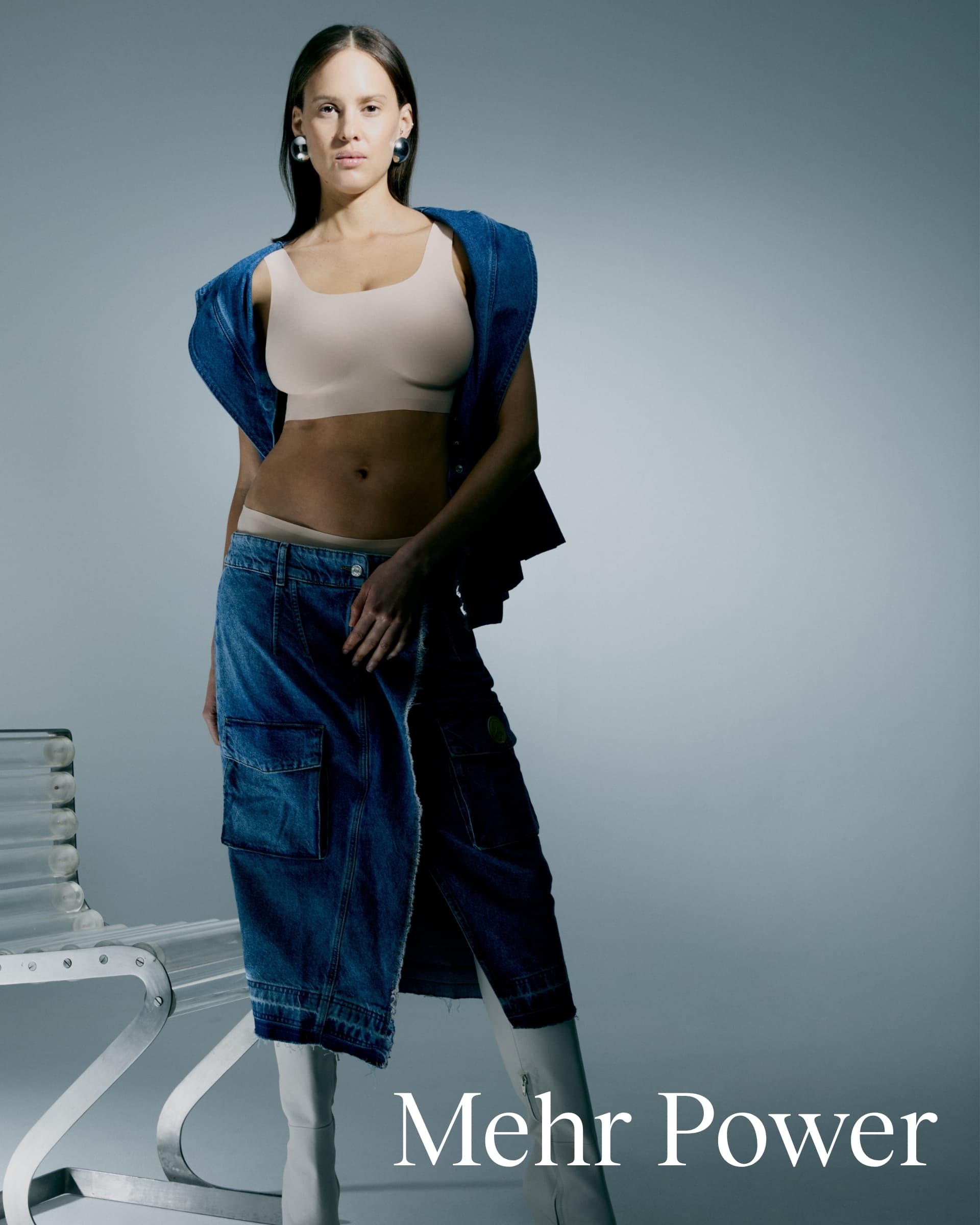 A woman in a nude bra and denim skirt poses confidently with a denim jacket over shoulders. Text reads "Mehr Power."