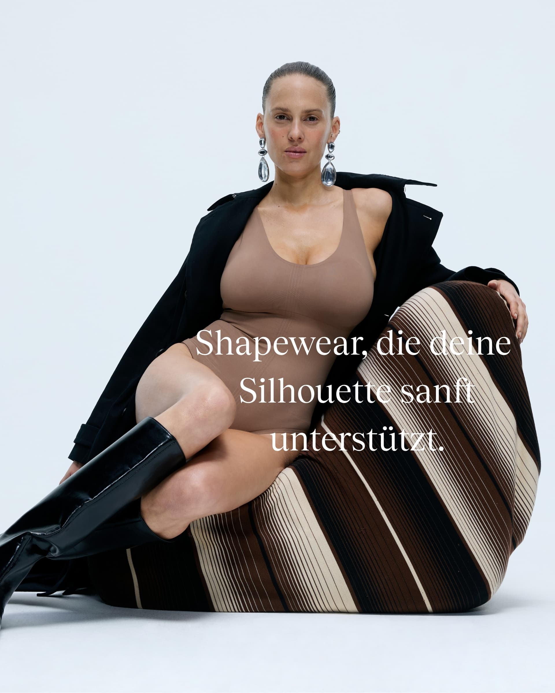 A woman poses in a brown and black outfit with bold earrings and knee-high boots, exuding confidence. Text reads: "Shapewear, die deine Silhouette sanft unterstützt."