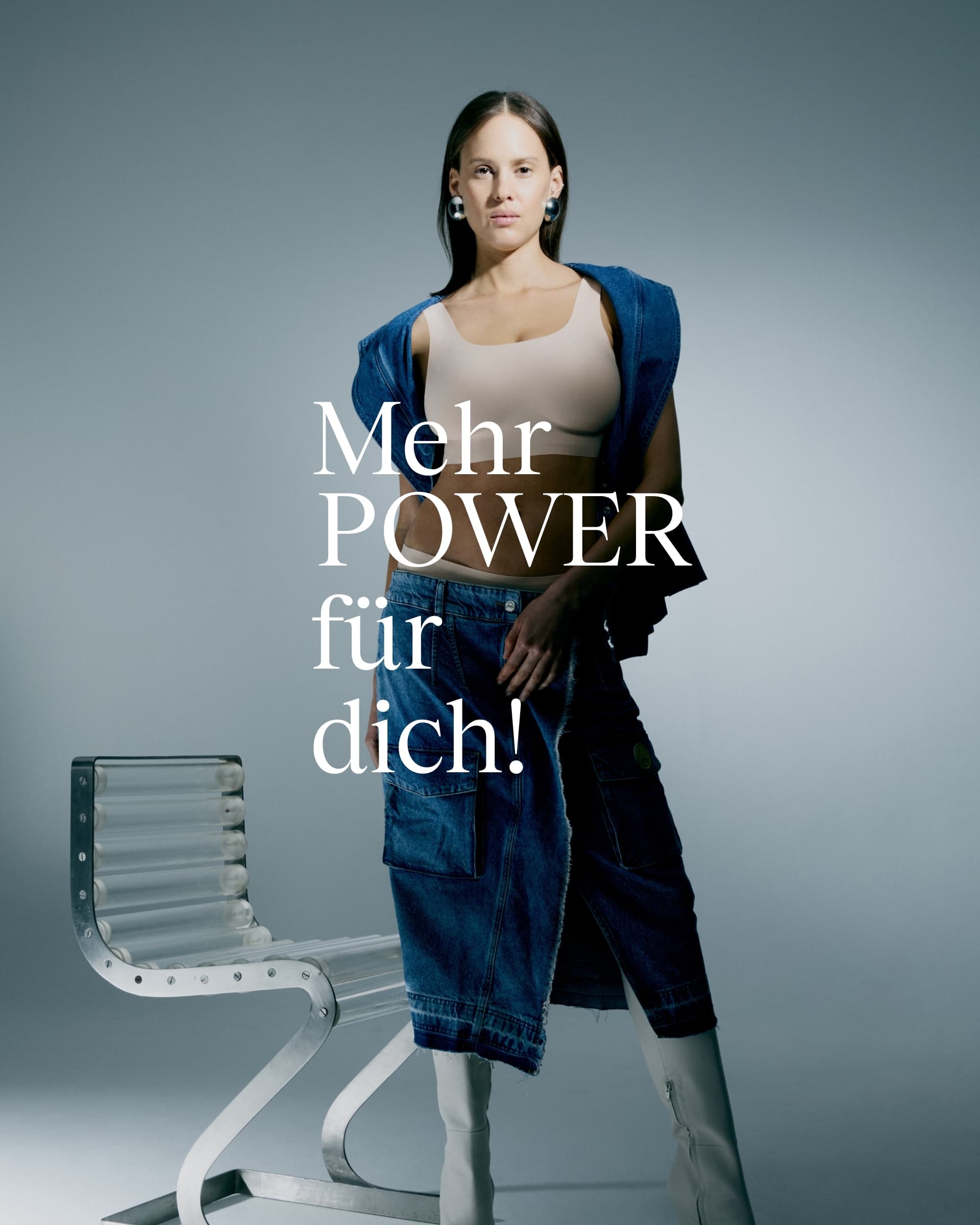 Woman in a denim outfit and boots stands confidently beside a modern chair, with text "Mehr POWER für dich!" in German.