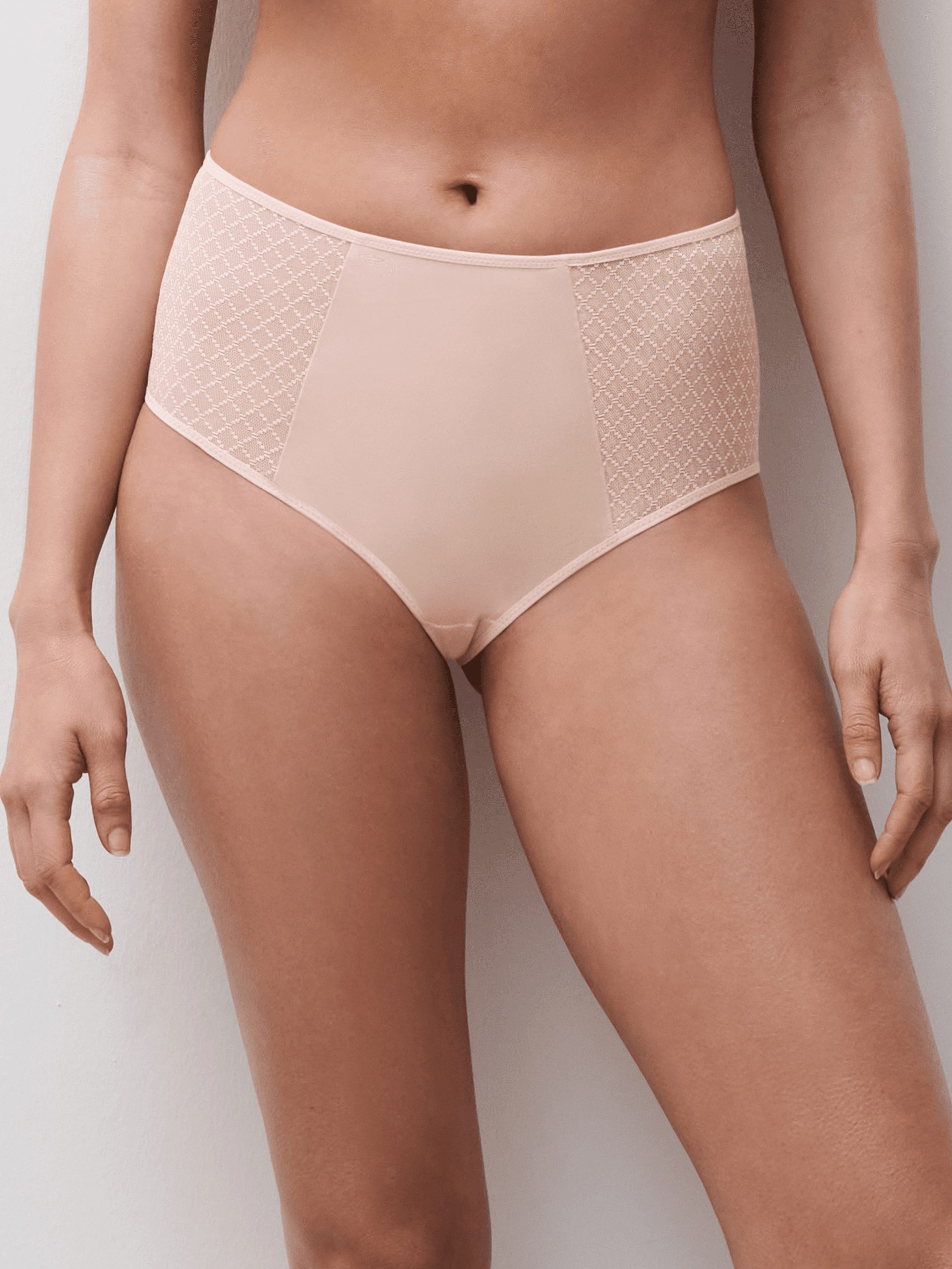 A person wearing high-waisted nude underwear with lace detailing on the sides, standing against a plain white background.