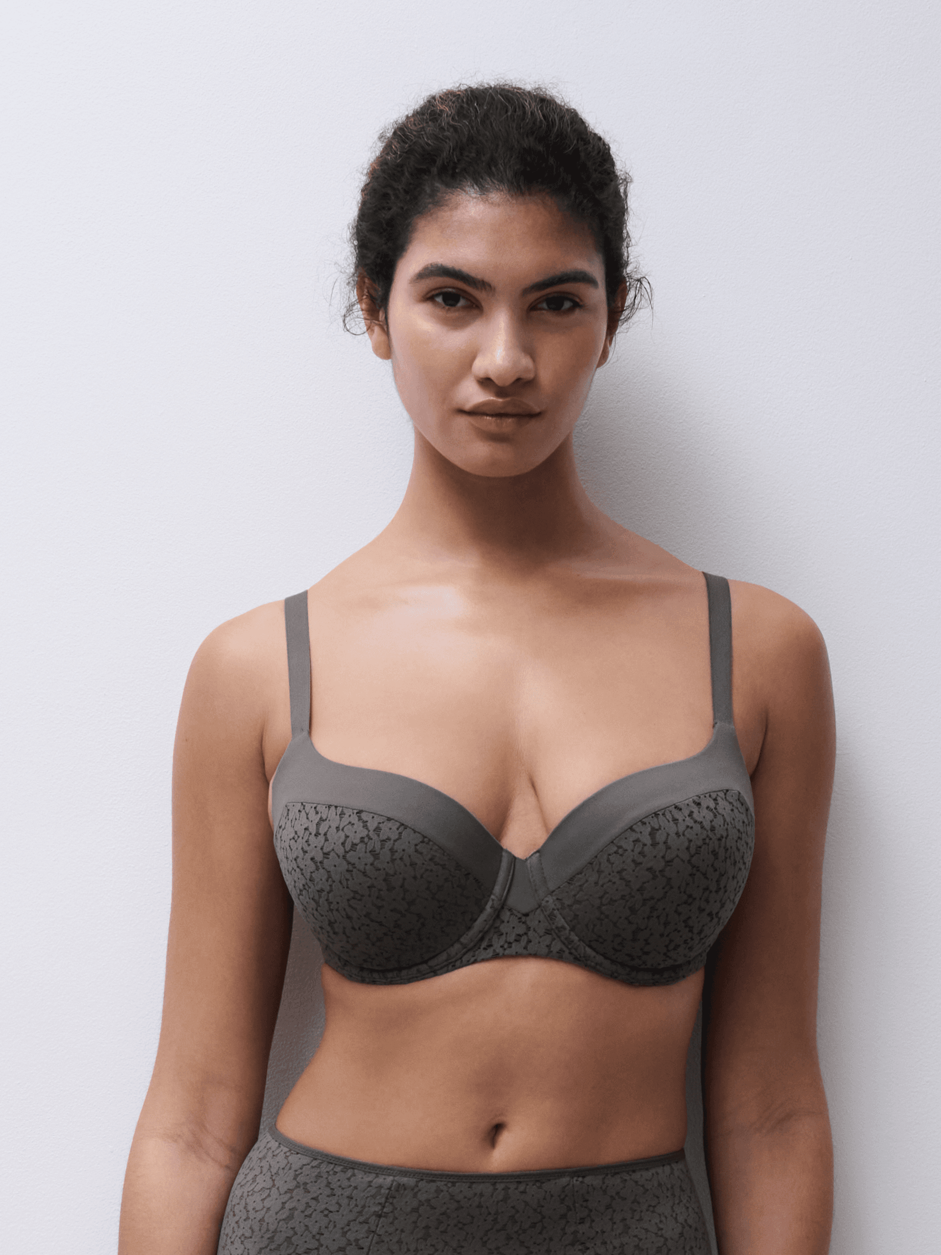 A woman wearing a green patterned bra stands against a plain white wall.